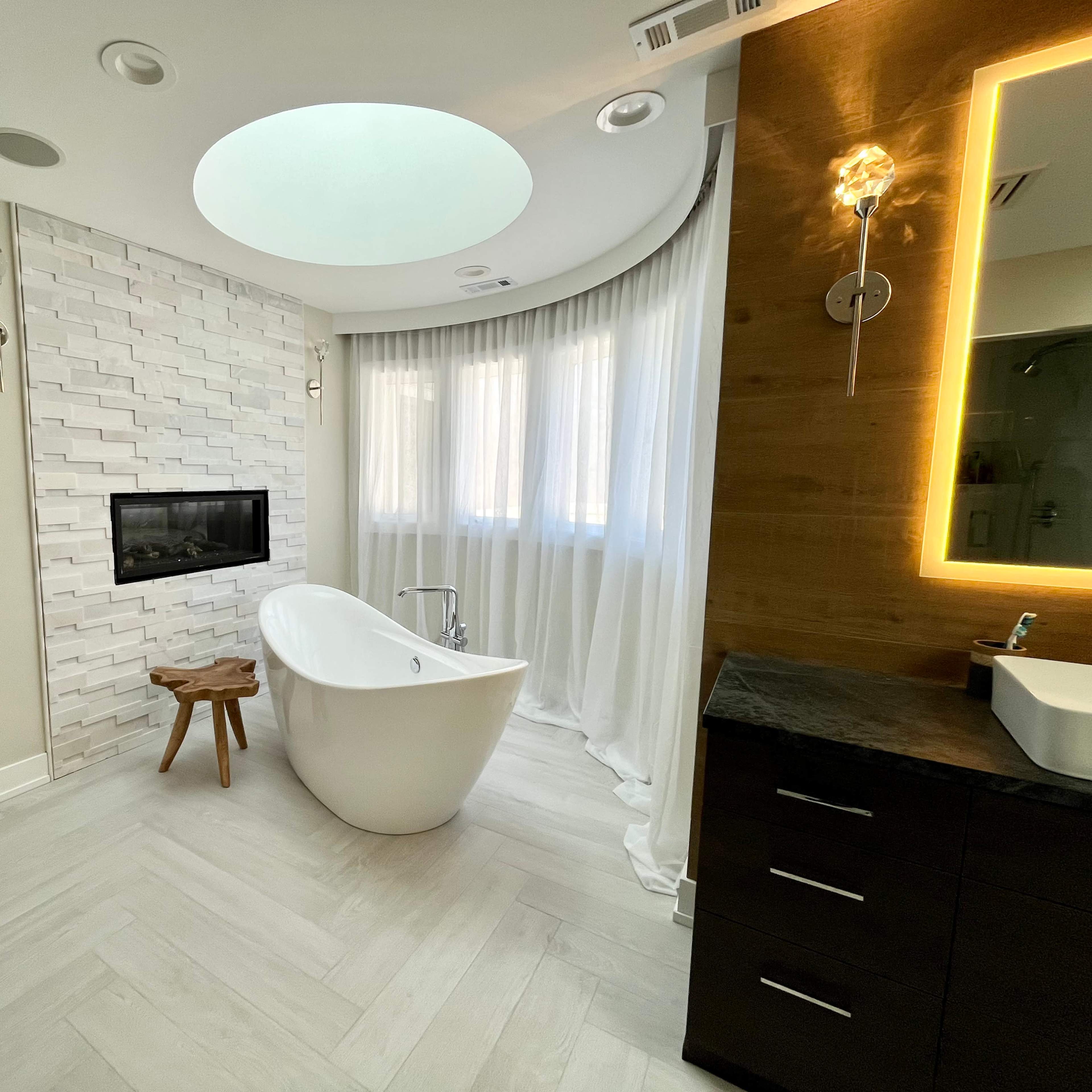 A modern bathroom features a freestanding tub, a wall-mounted fireplace, and large windows with sheer curtains.