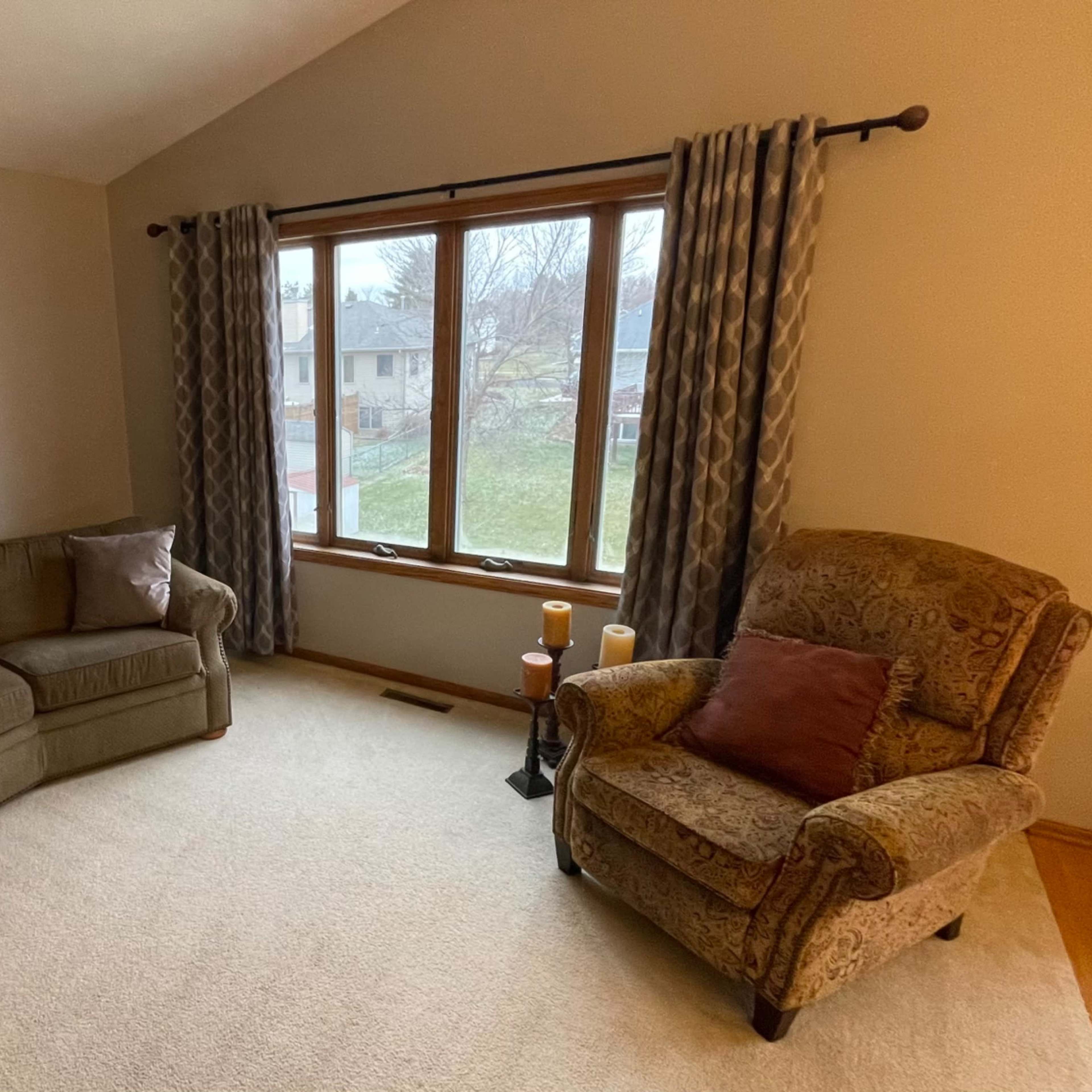 A cozy living room features a curved sofa, an armchair, and large windows with drapes overlooking a yard.