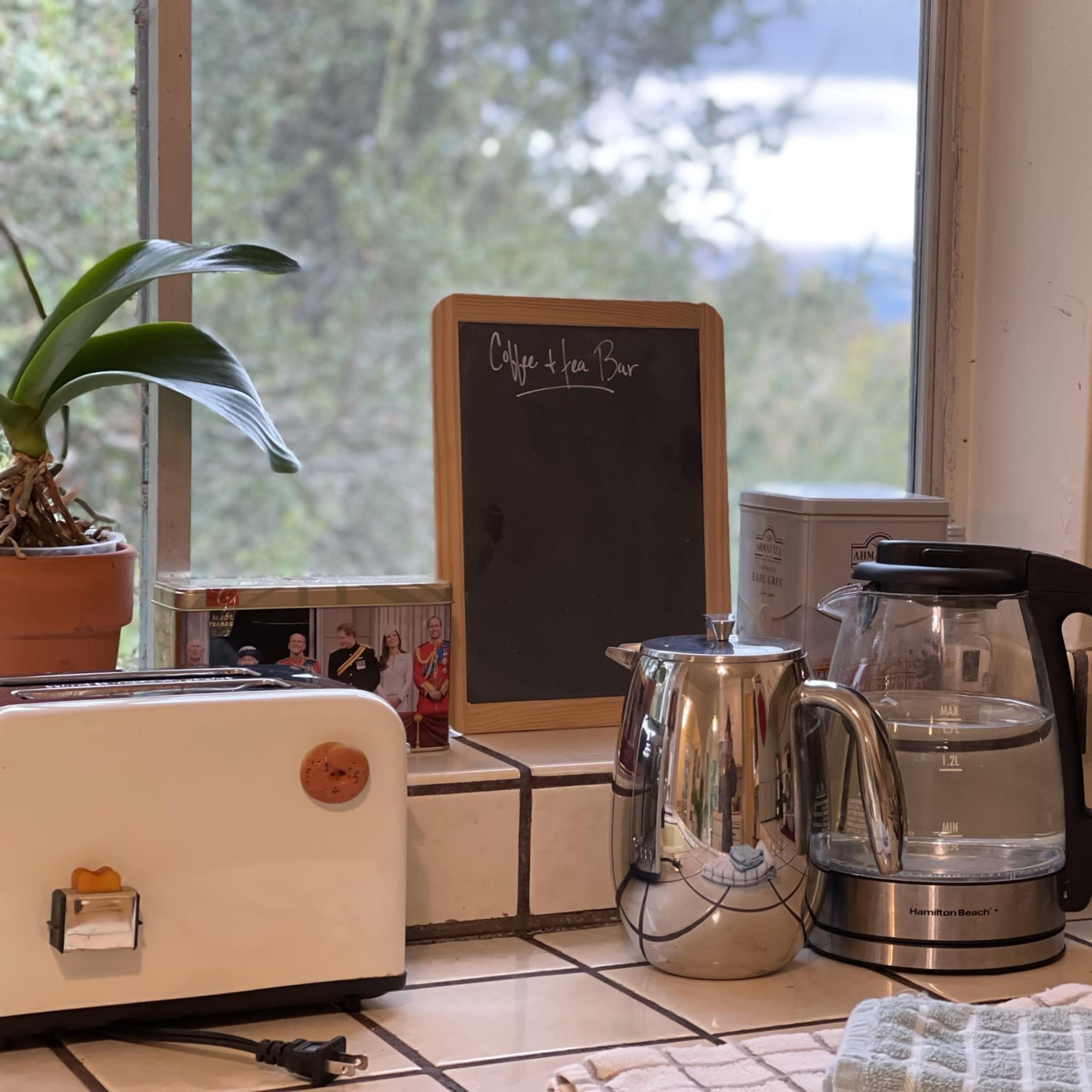 A kitchen counter with a toaster, a coffee maker, a kettle, and a chalkboard, along with a potted plant by the window.