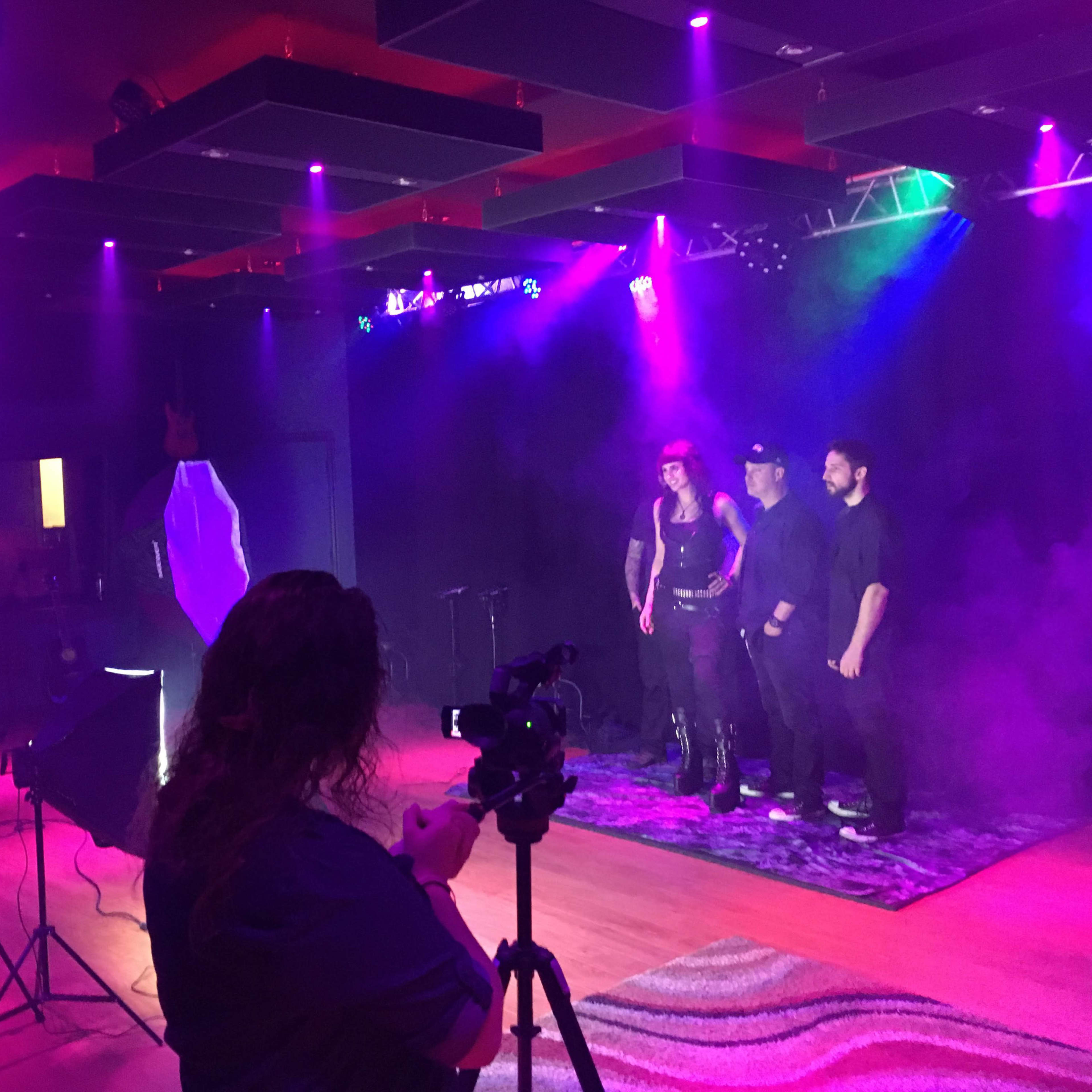 A group of three people poses on stage under purple lighting, while a person captures the moment with a camera.
