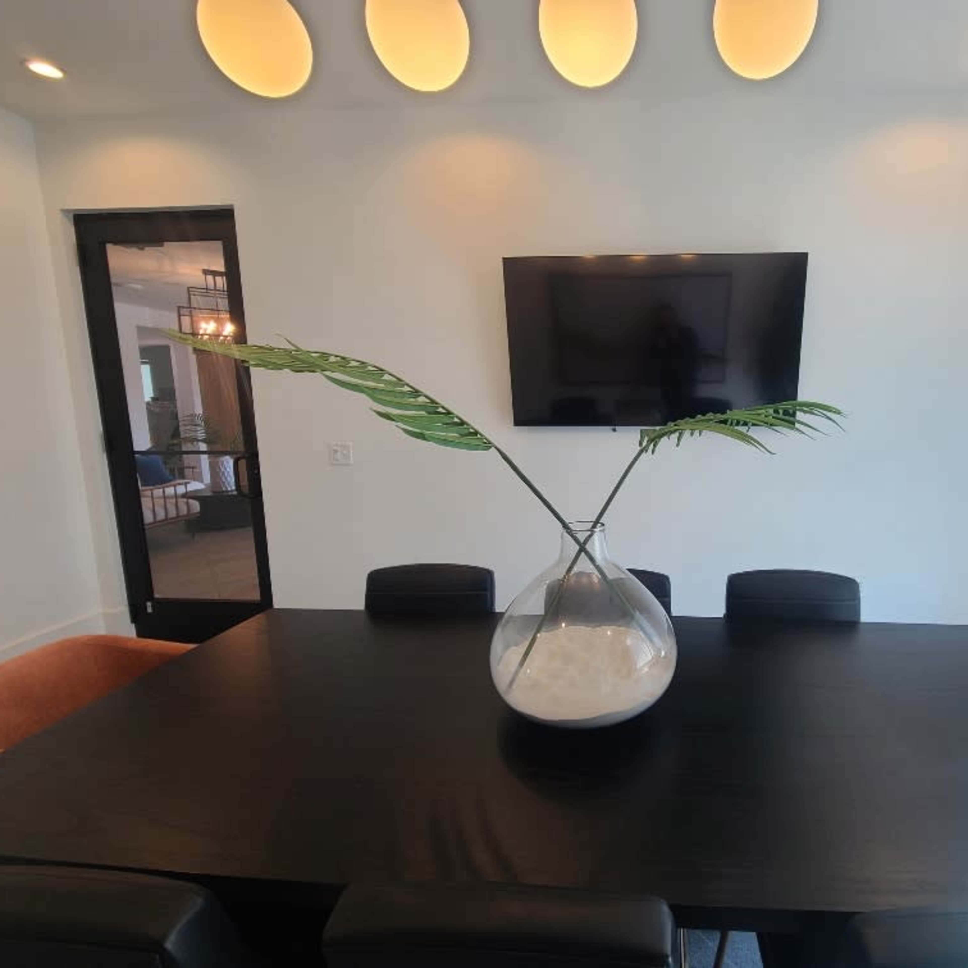 A modern dining room features a black table with a large glass vase containing a decorative plant, complemented by a wall-mounted television and illuminated ceiling lights.