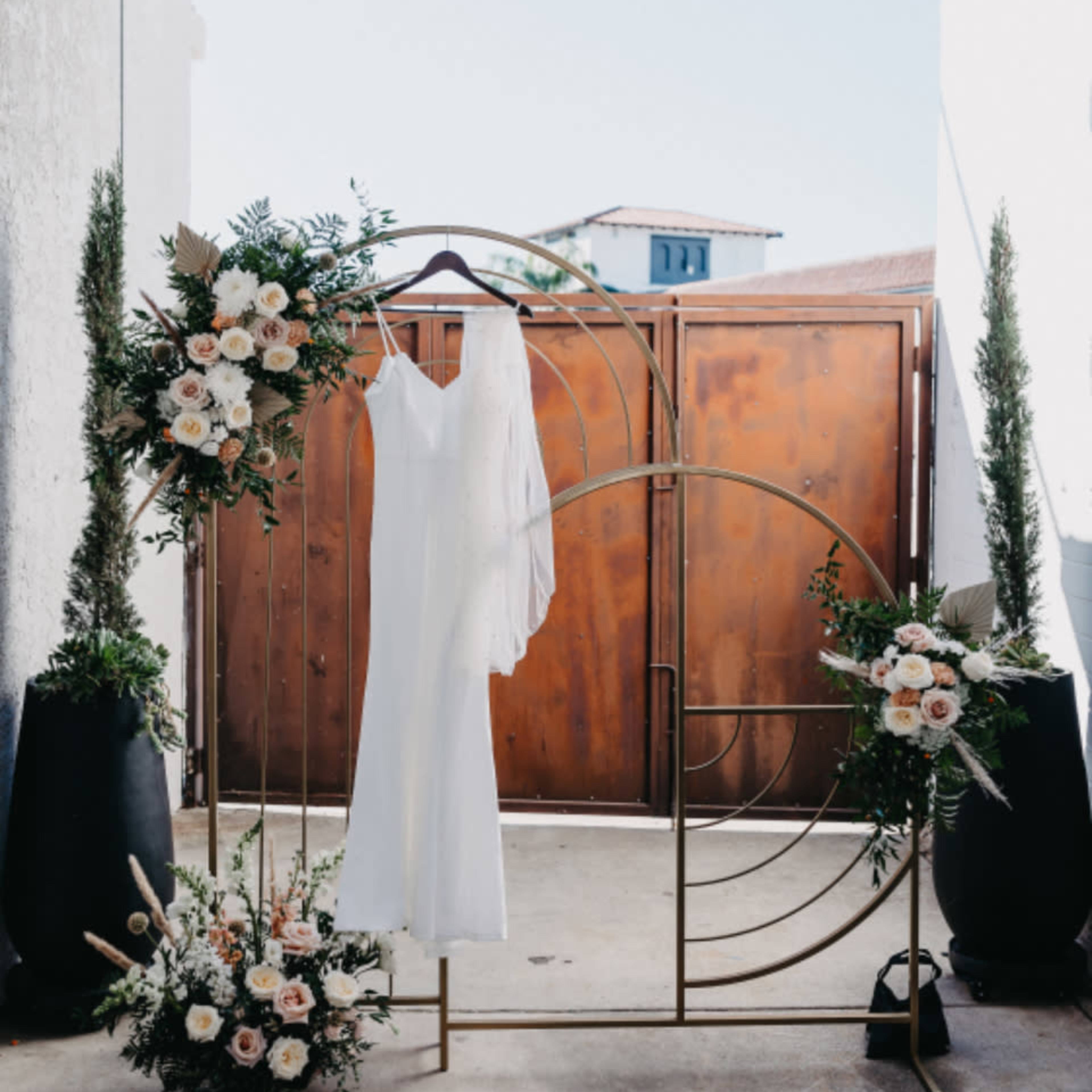A white wedding dress is displayed on a hanger in front of a decorative backdrop made of gold geometric shapes and surrounded by floral arrangements.