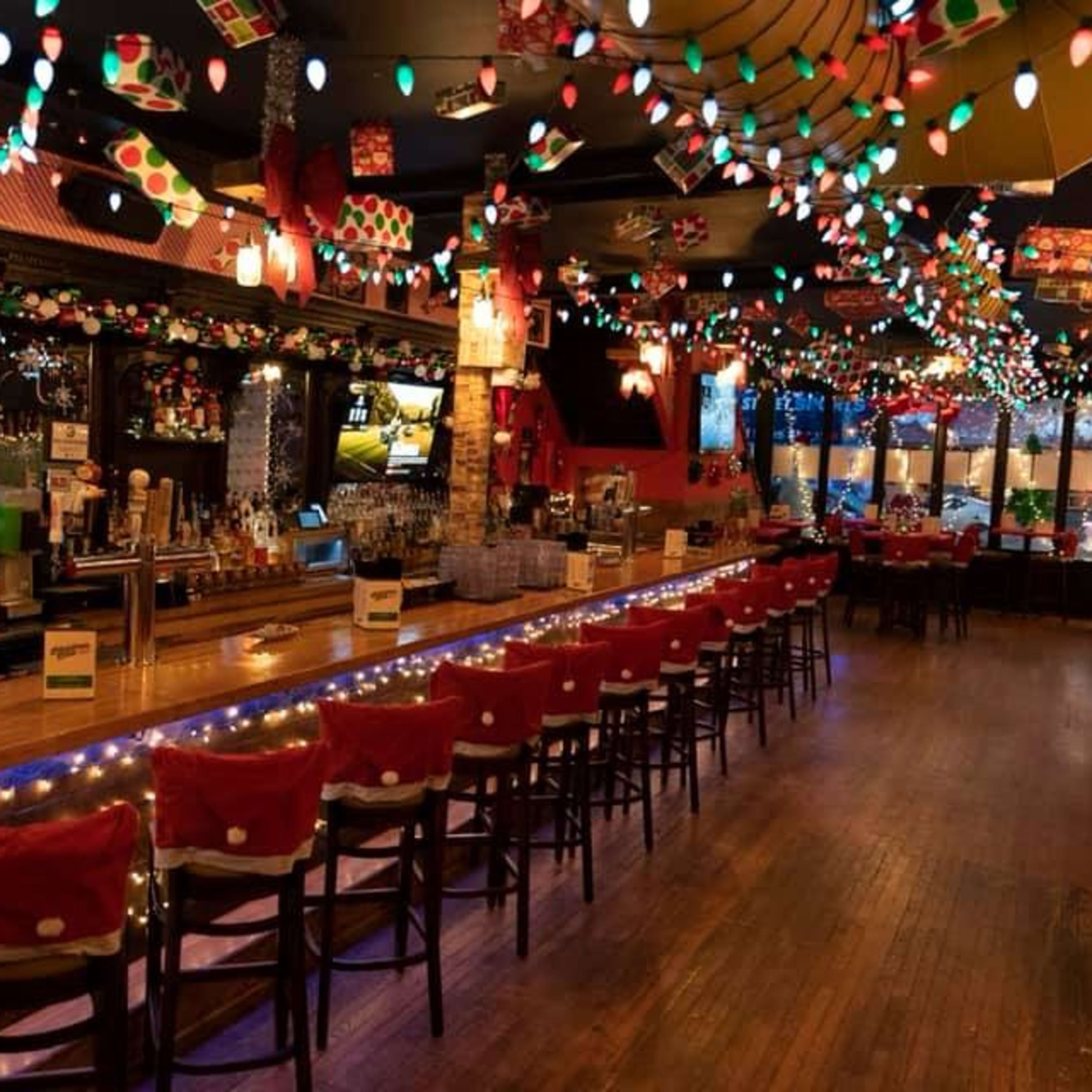 A bar adorned with holiday decorations, featuring a long wooden counter and colorful string lights hanging from the ceiling.