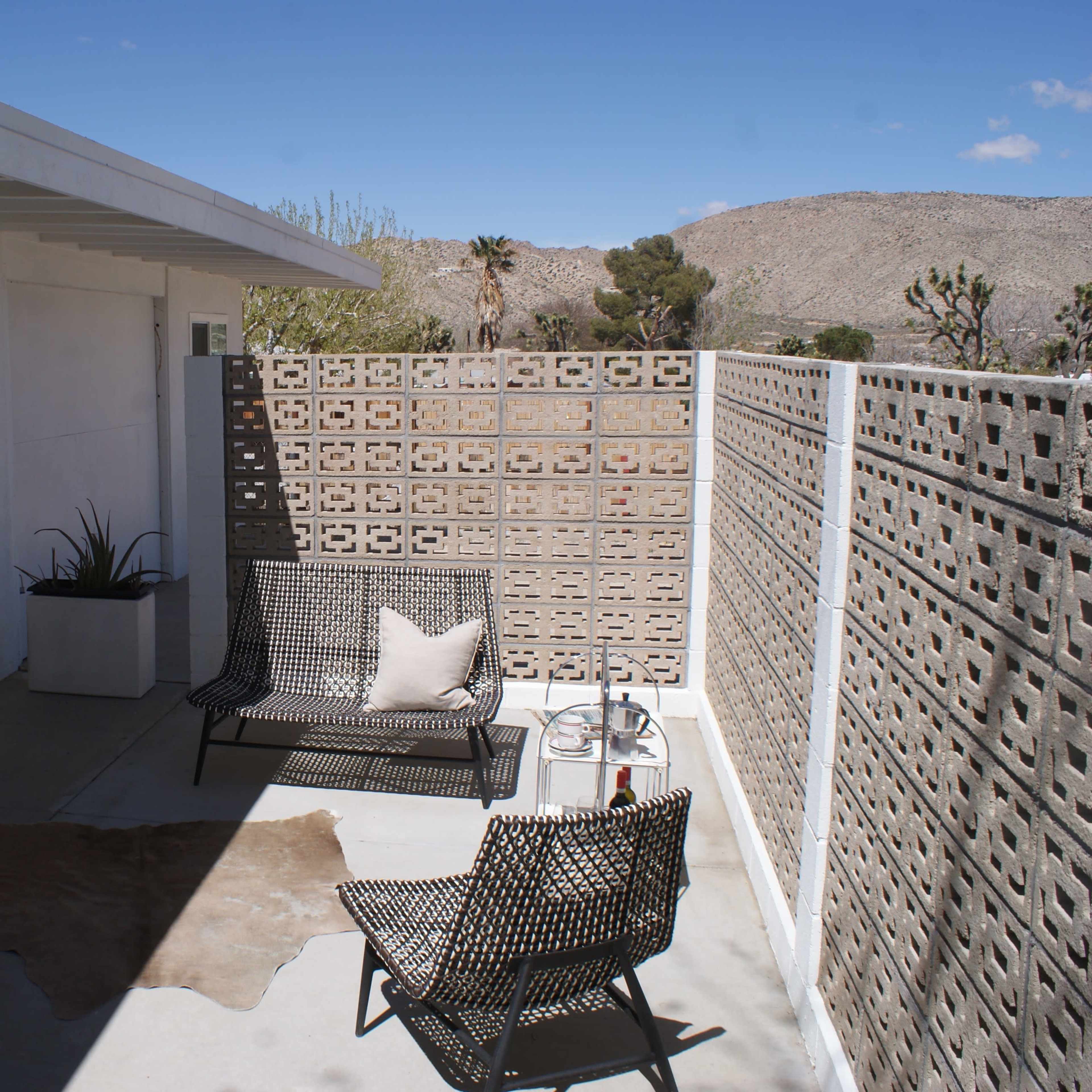 A small outdoor patio features angled seating, a potted plant, and decorative concrete block walls against a backdrop of desert hills and blue sky.