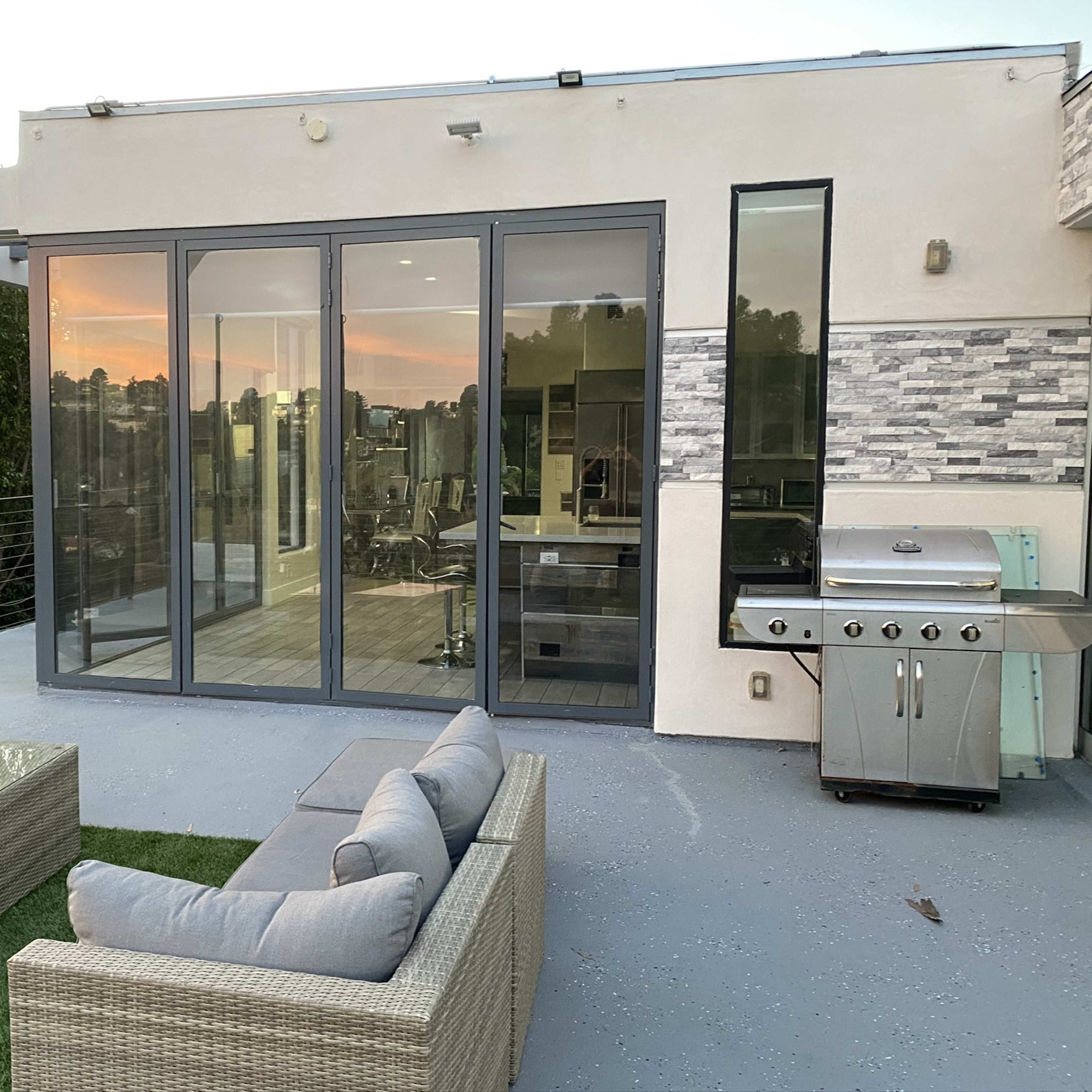 A modern patio area with outdoor seating, a glass door opening to an interior space, and a barbecue grill, set against a backdrop of trees and a colorful sunset.