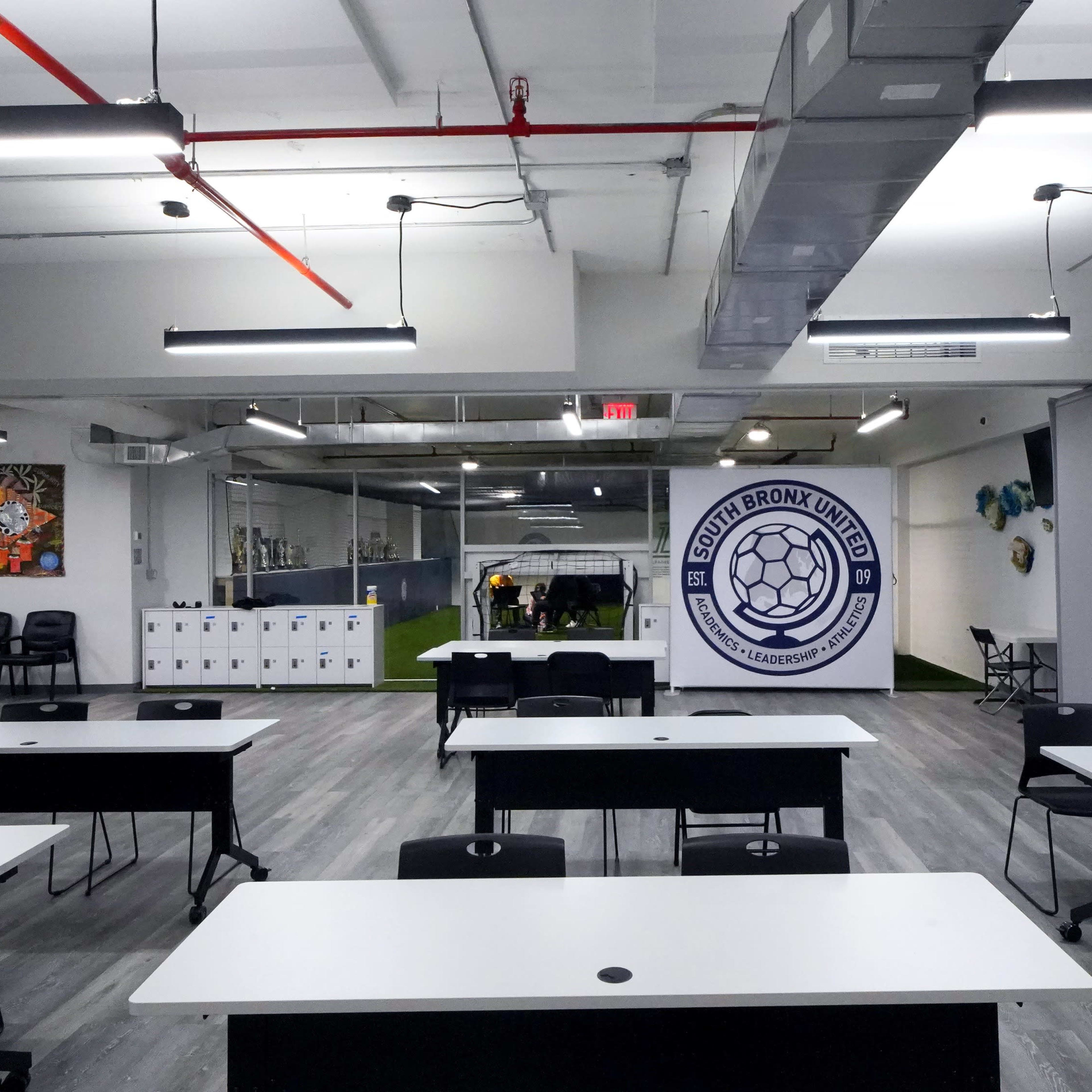 A modern classroom or meeting space with multiple tables and chairs arranged in rows, featuring a large logo of a soccer team on the wall.