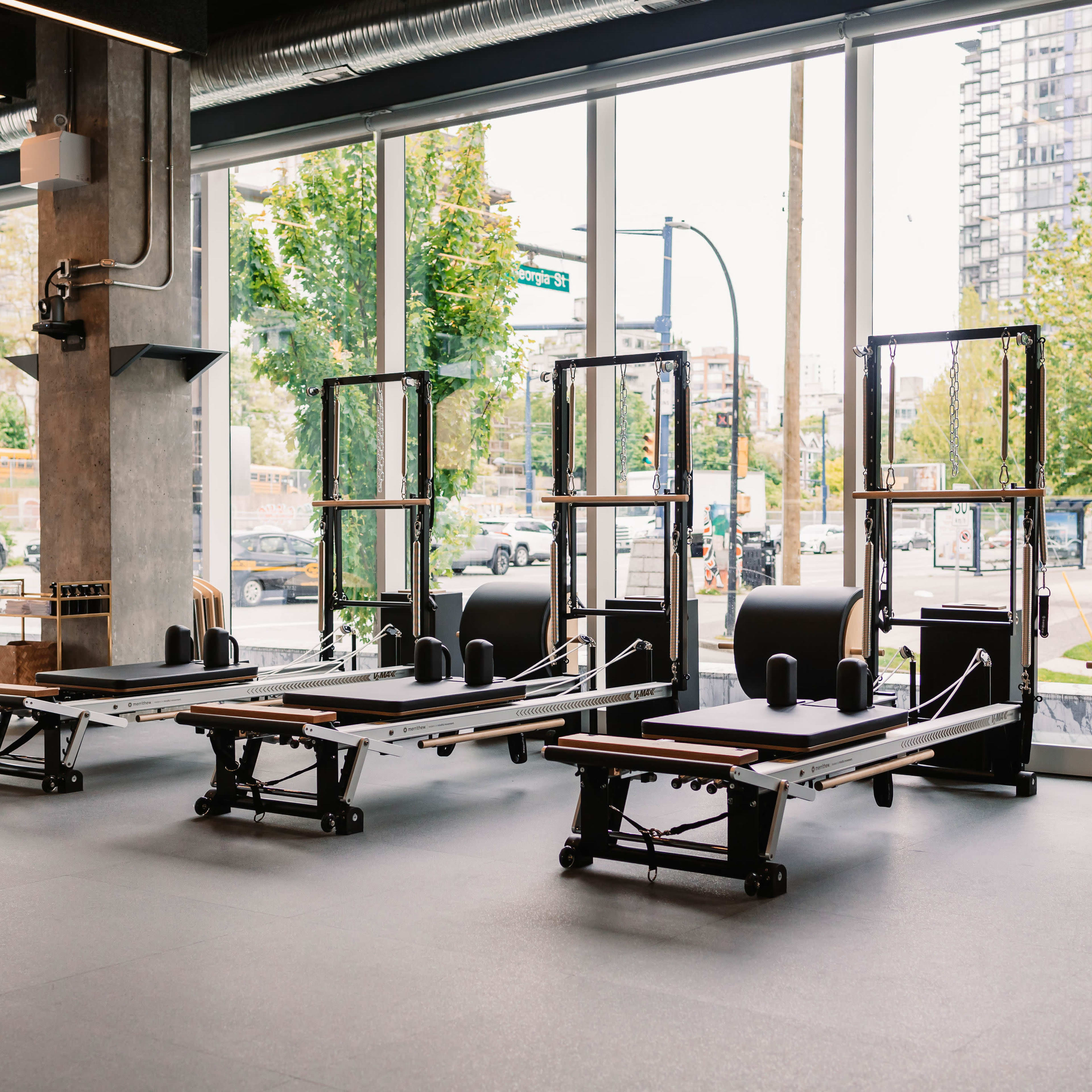 A bright fitness studio featuring several reformer Pilates machines set against large windows.