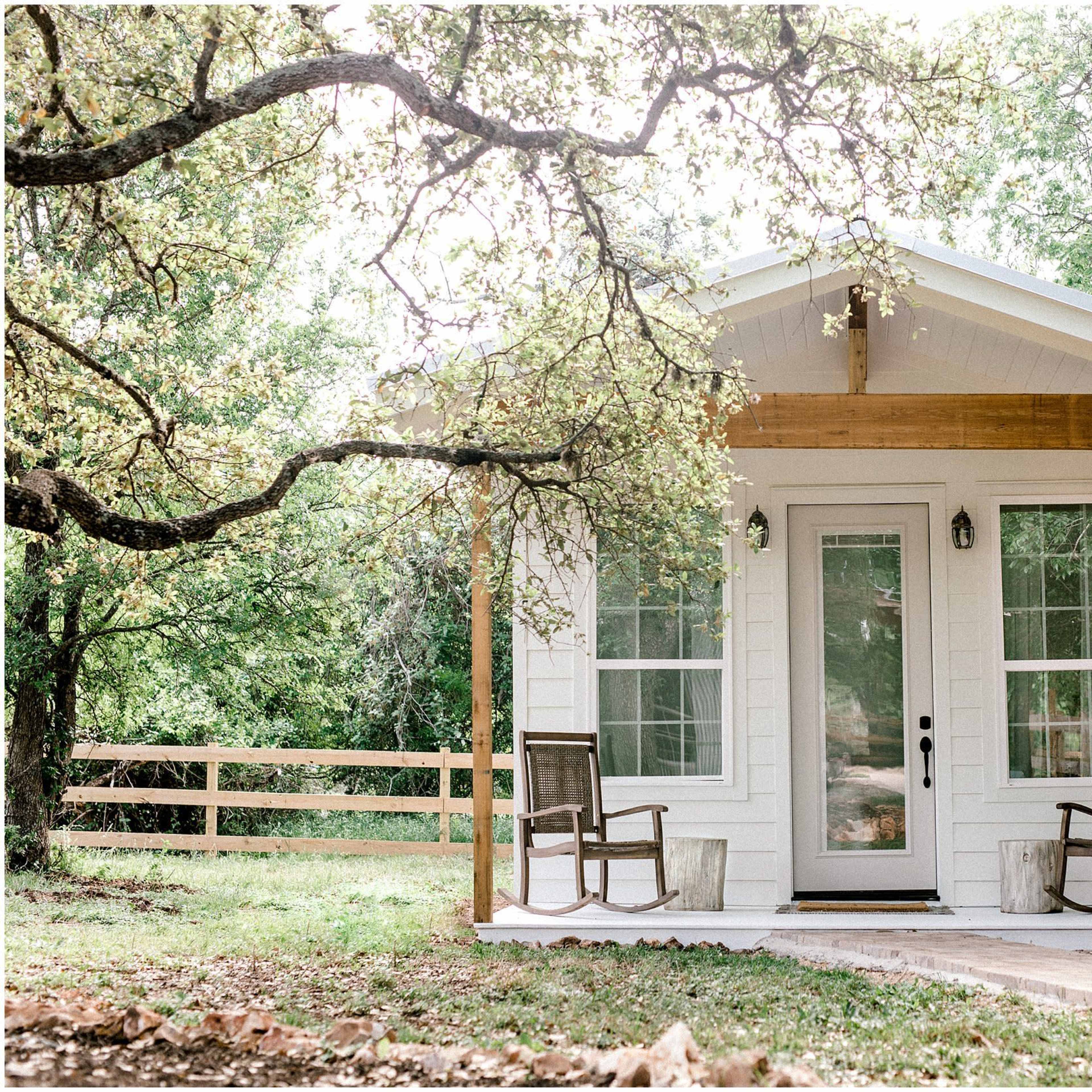 A small white cottage with a front porch and two wooden chairs is surrounded by trees and a wooden fence.