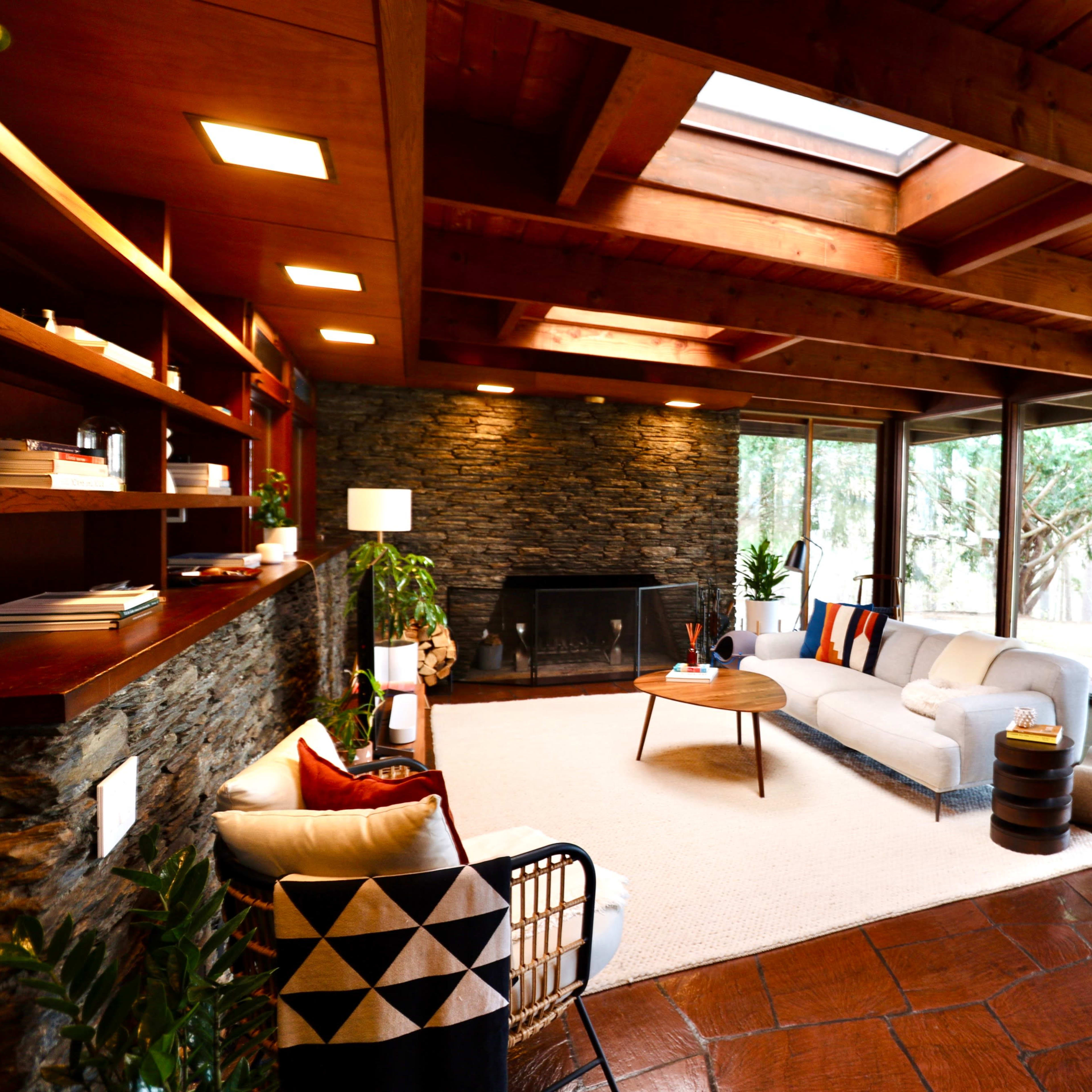 A spacious living room with wooden beams, a stone wall, and large windows overlooking a forested area.