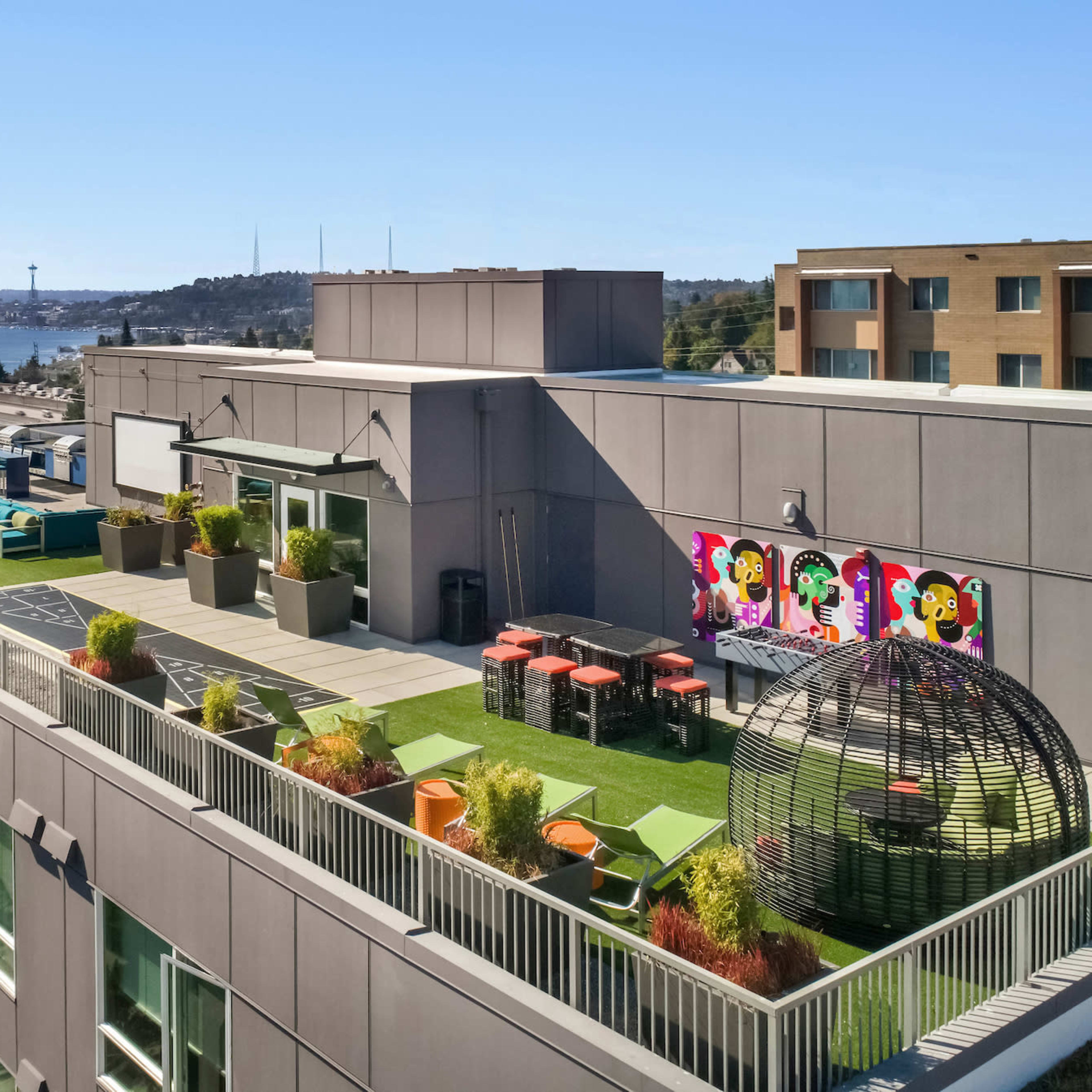 A rooftop garden with seating areas, colorful artwork on the wall, and a view of a city skyline in the background.