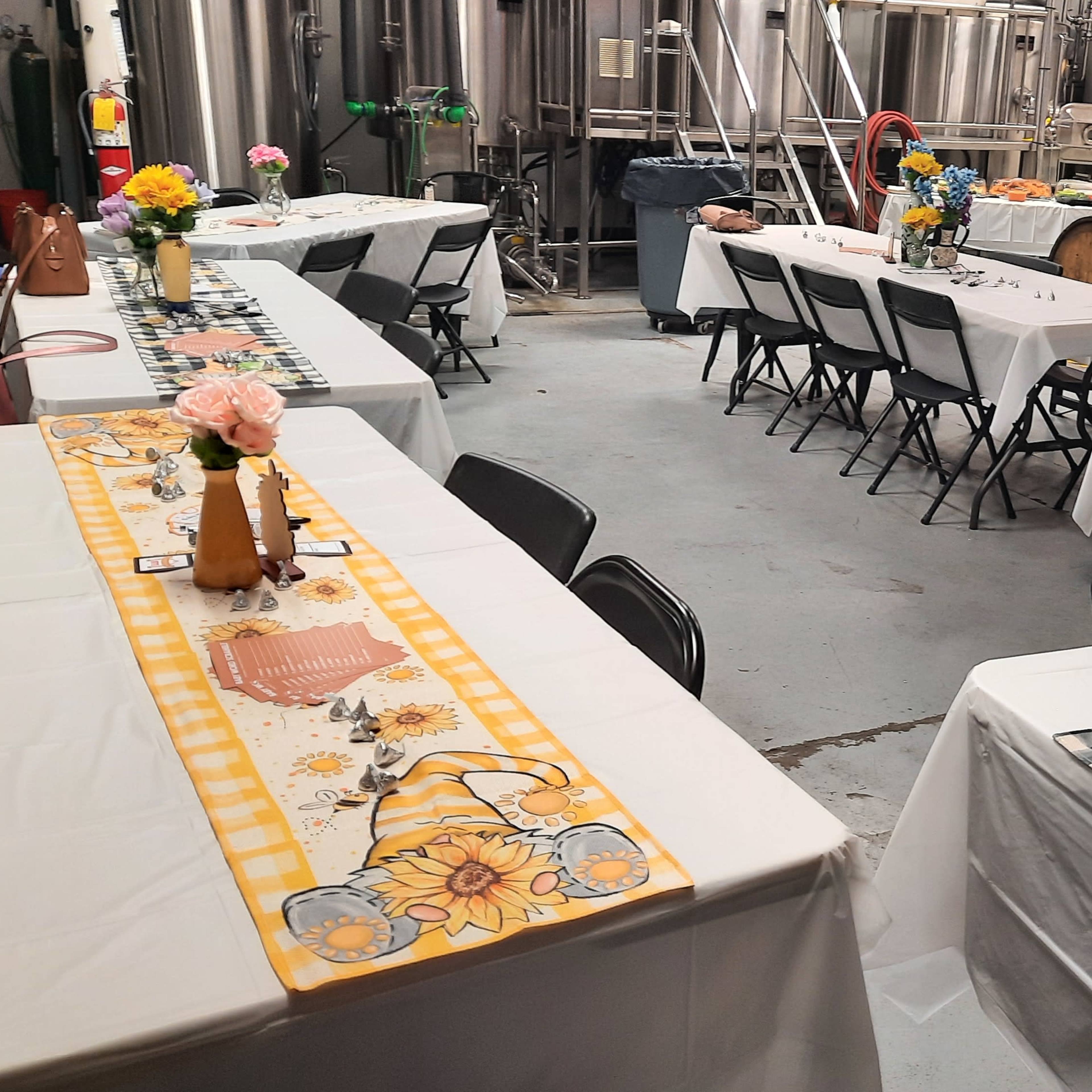 A room set up for an event, featuring several tables covered with white tablecloths, decorated with floral centerpieces, and surrounded by black chairs, with brewing equipment visible in the background.