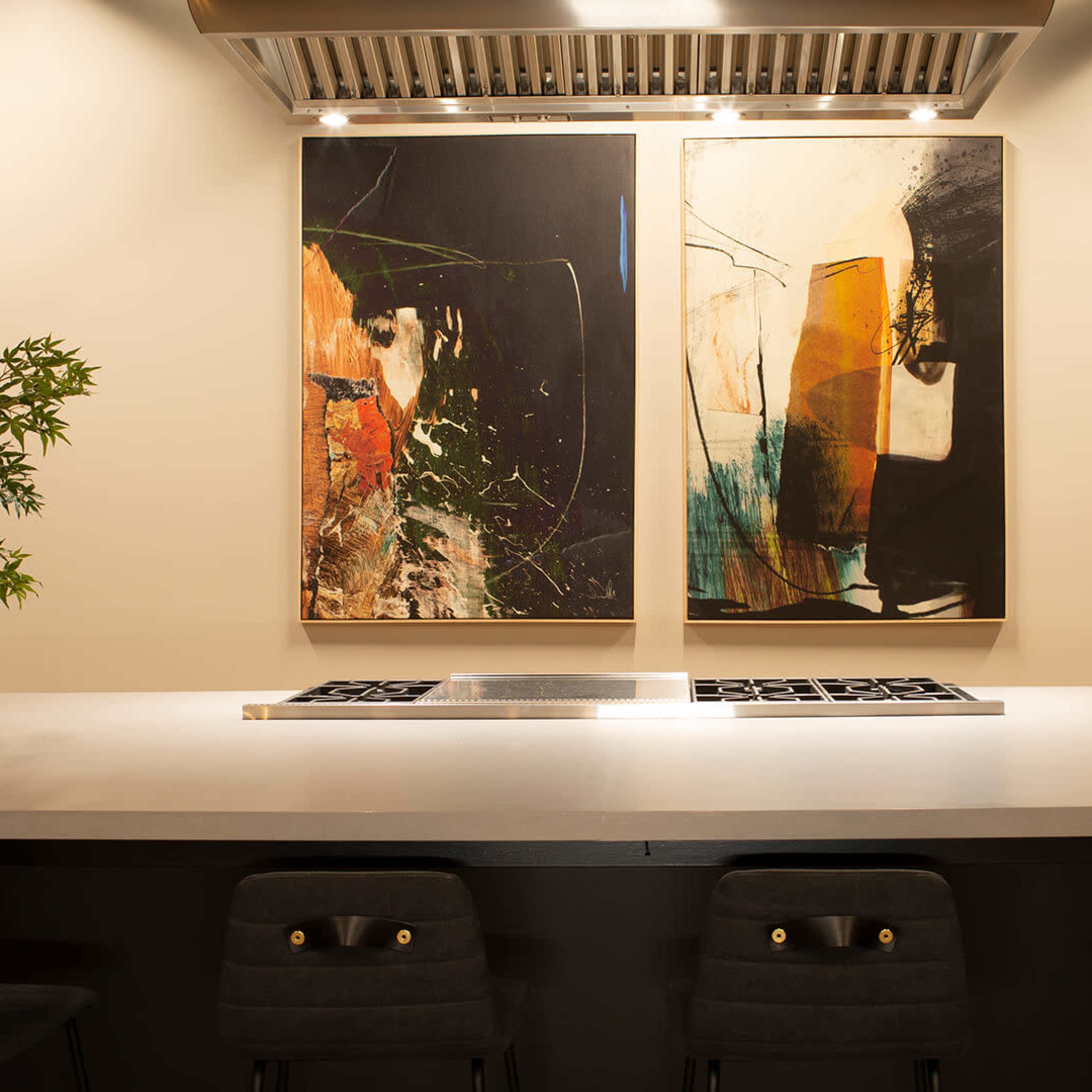 A modern kitchen features a large island with a cooktop and four barstools, complemented by two abstract paintings on the wall.