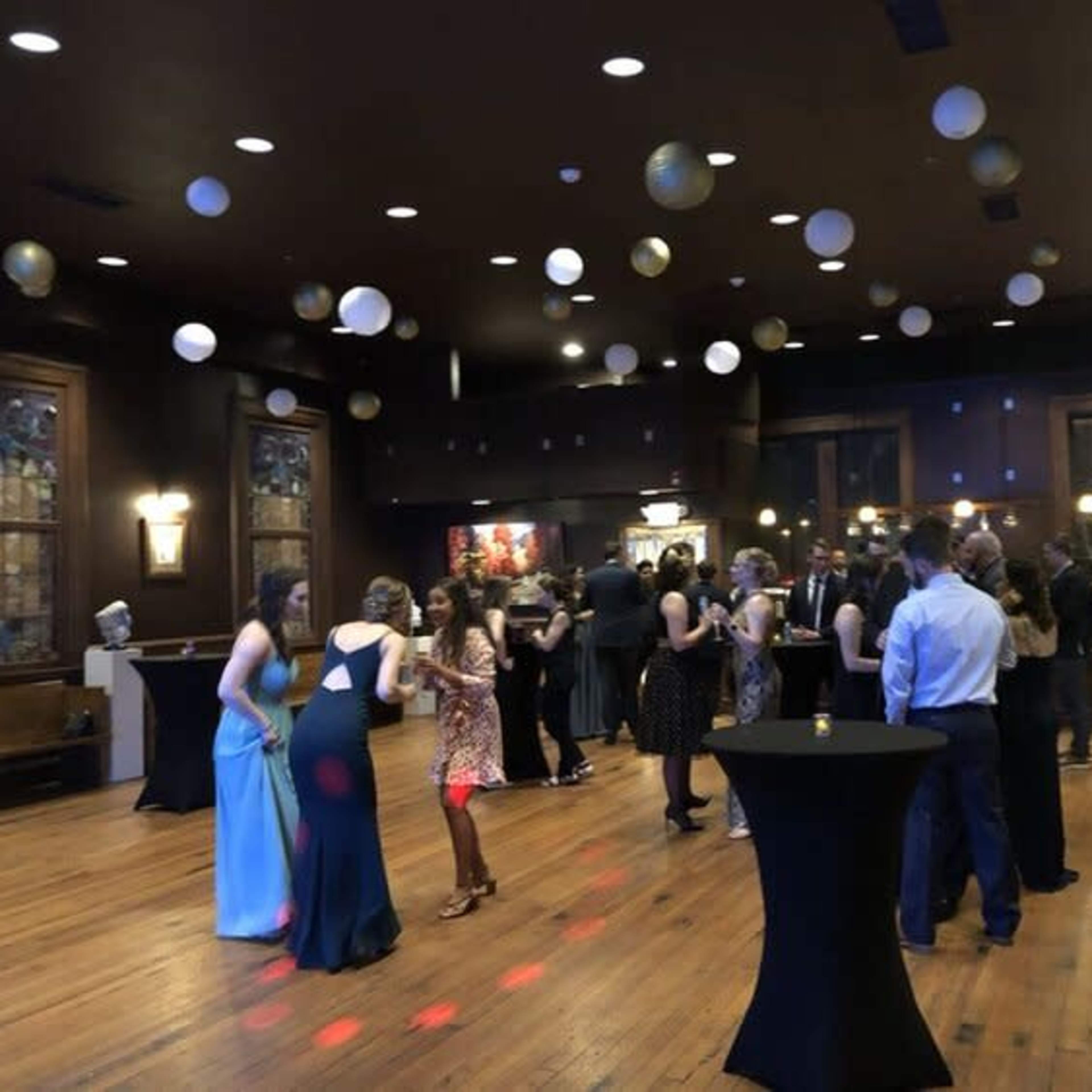 A lively gathering takes place in a decorated hall with people dancing and standing around tables under hanging decorative lights.