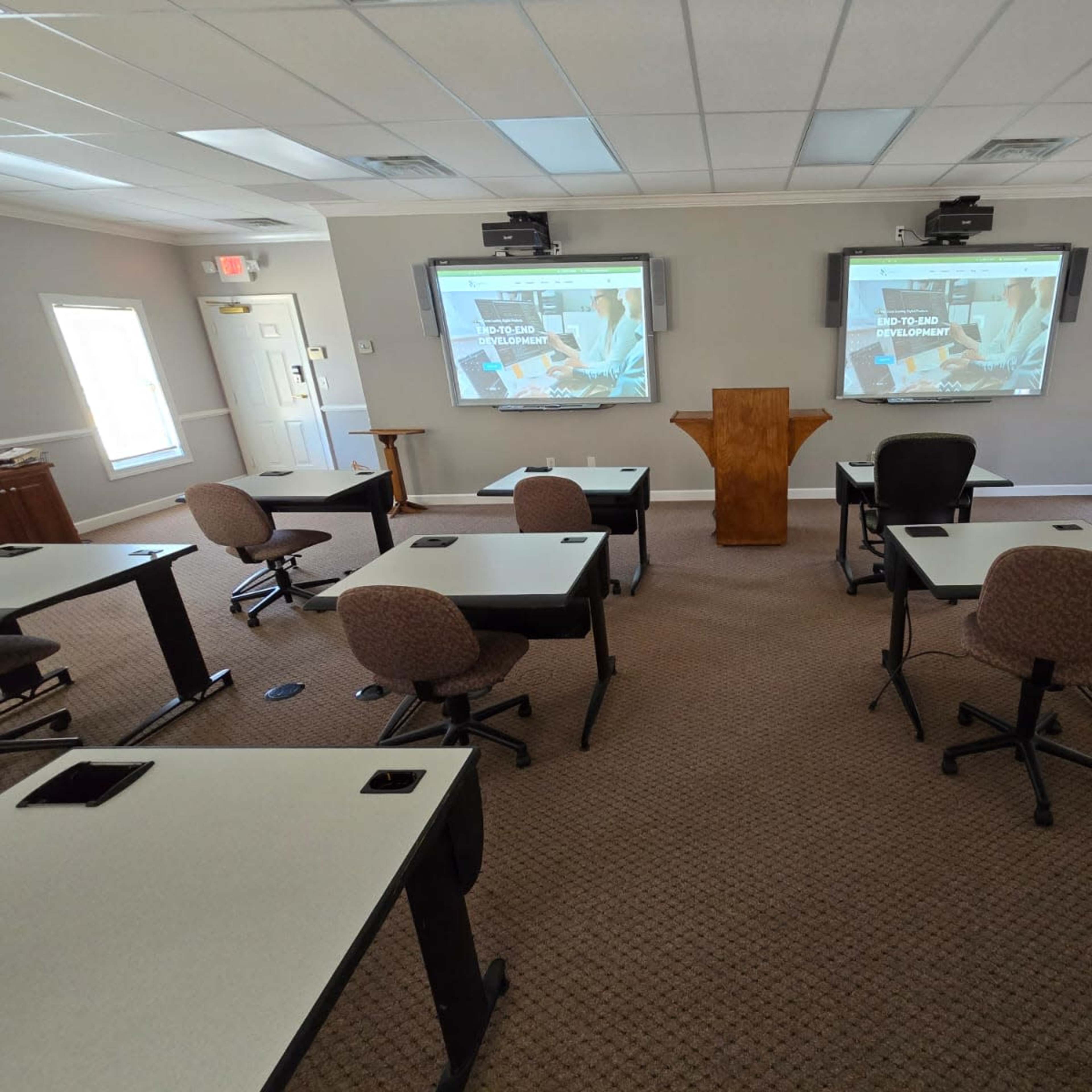 A classroom is set up with white tables and rolling chairs facing two large screens displaying presentation slides.