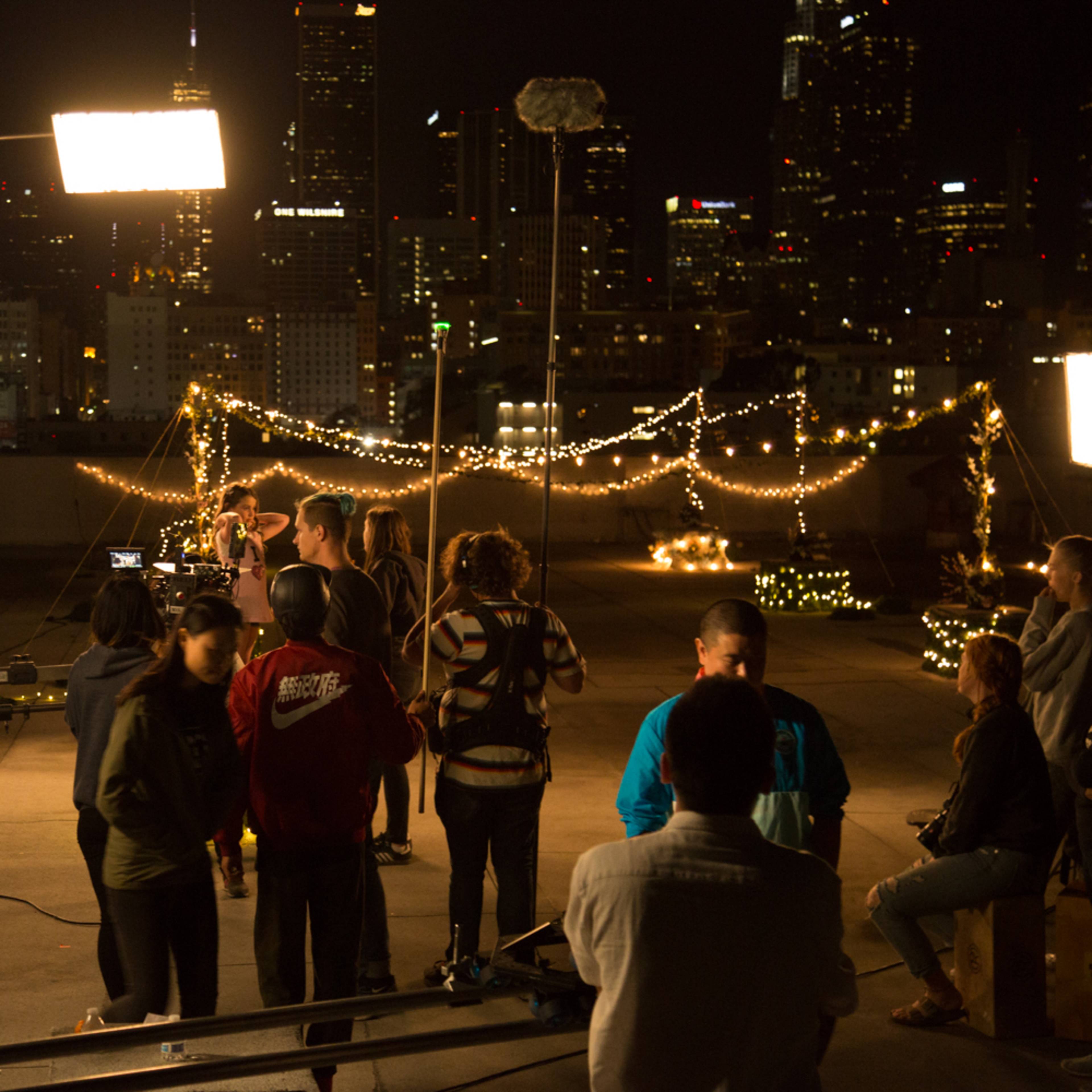 A film crew sets up equipment on a rooftop at night, with the city skyline illuminated in the background.