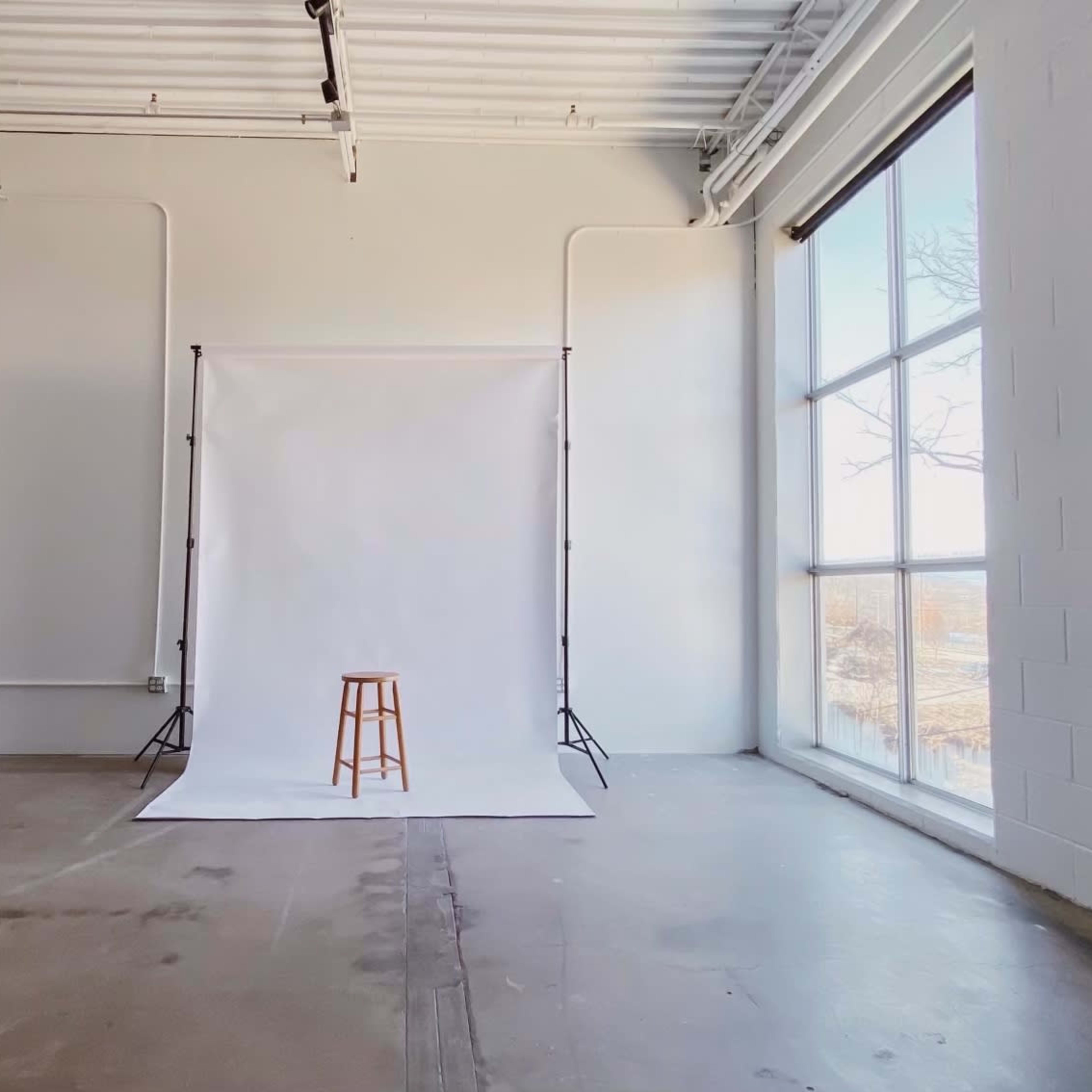 A spacious indoor studio with a plain white backdrop, a wooden stool positioned in front of it, and large windows allowing natural light to enter.