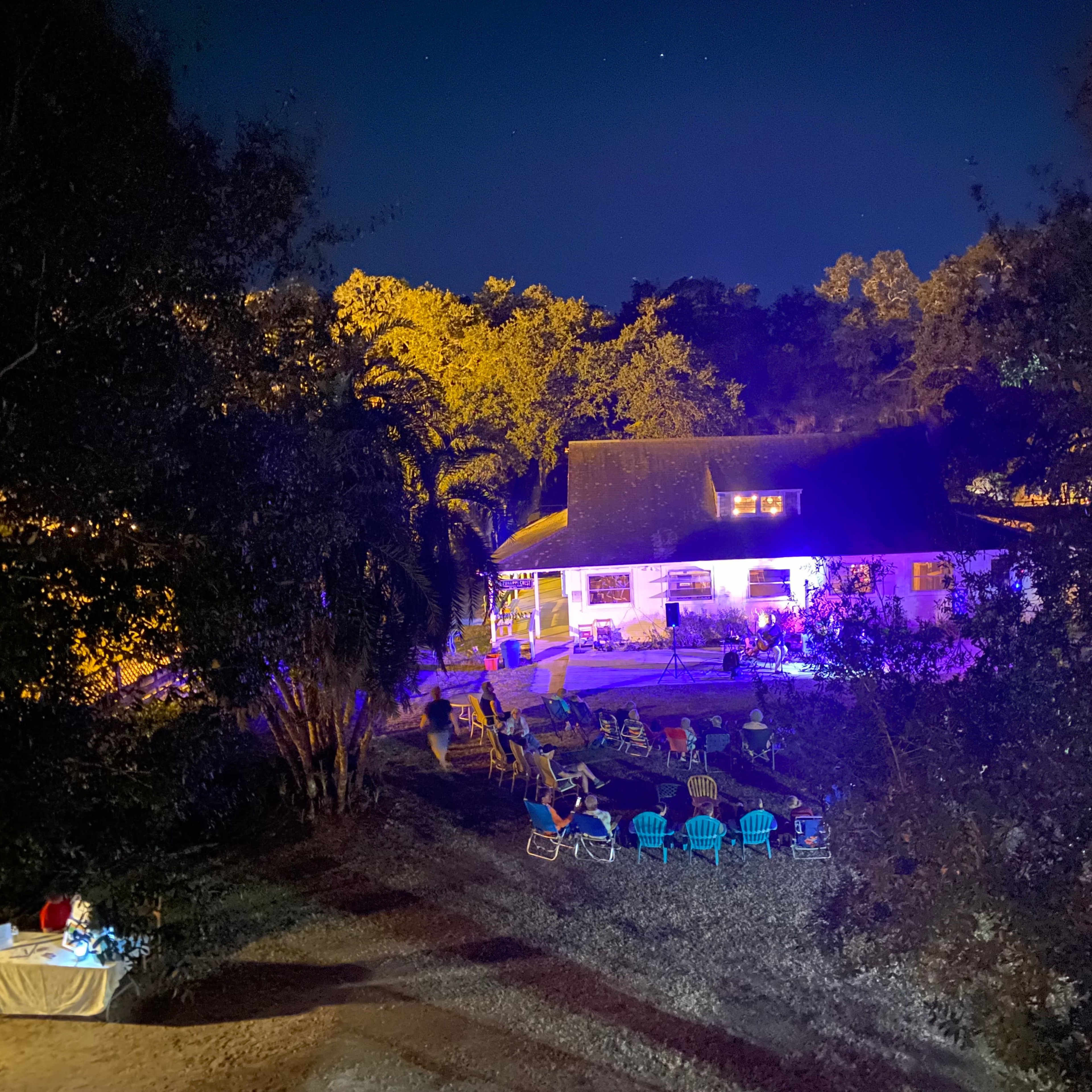 A house is illuminated by colorful lights at night, surrounded by chairs arranged on a grassy area amidst trees.