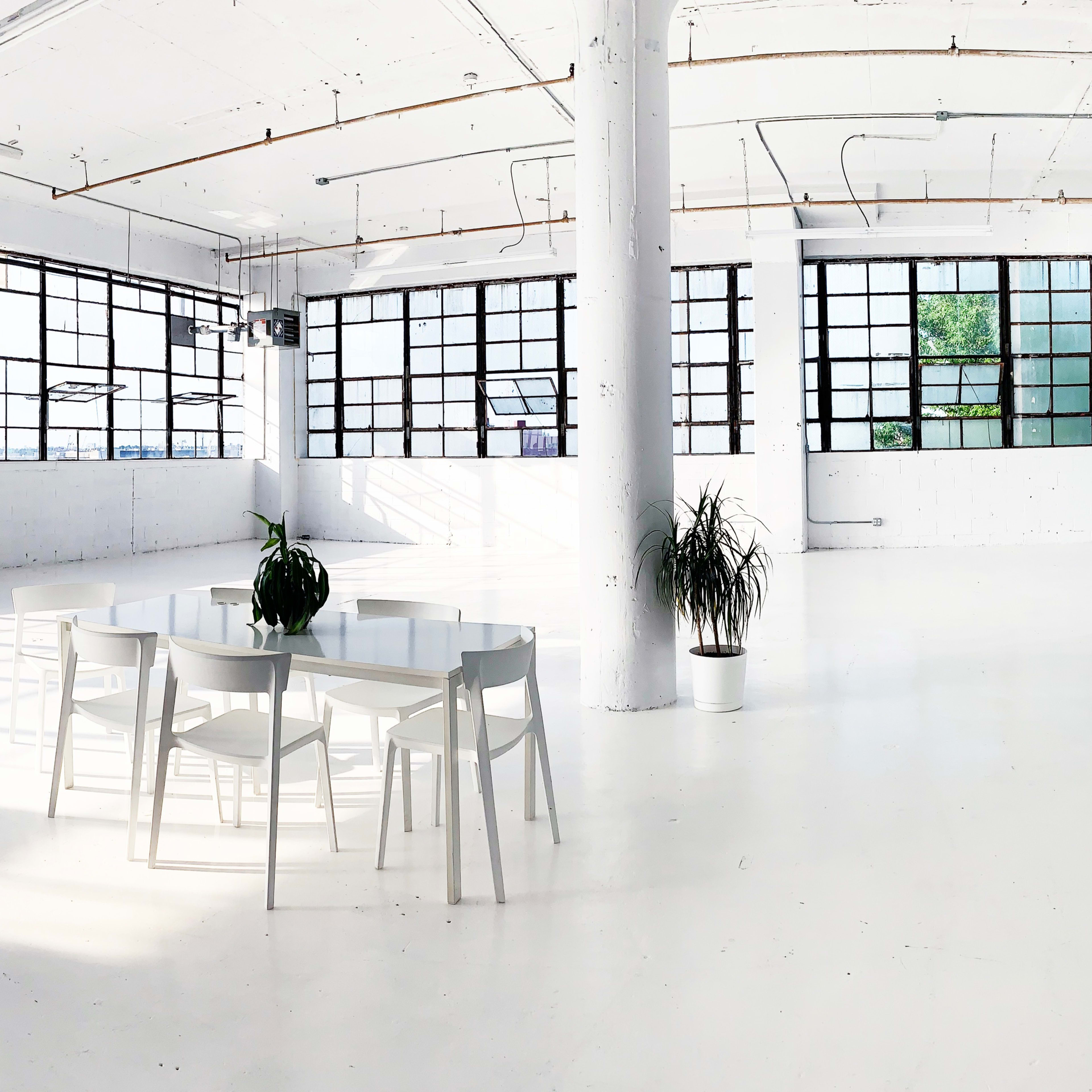 A spacious, brightly lit room with large windows, featuring a white circular table surrounded by chairs and potted plants.