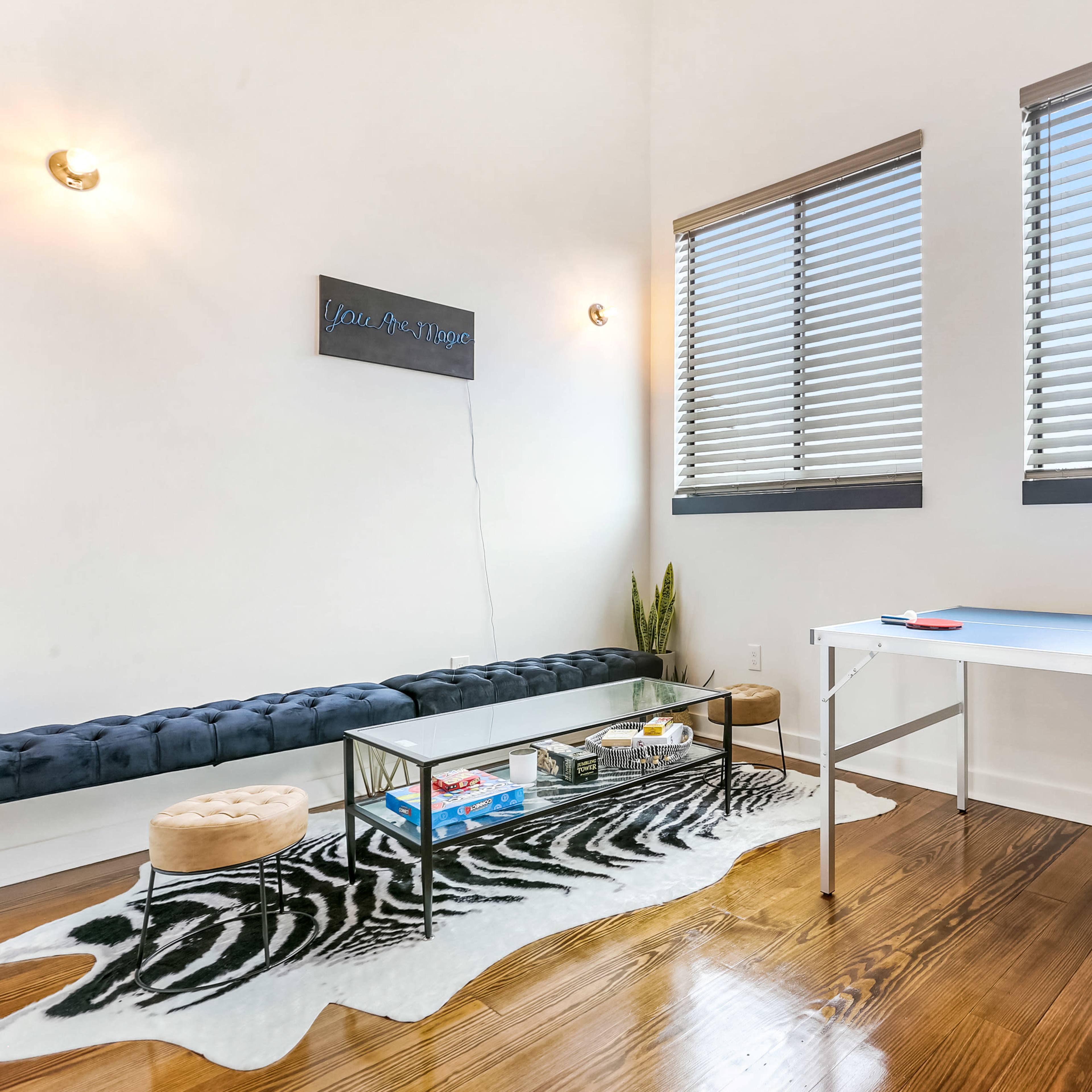 A room with a modern design featuring a black tufted bench, a glass coffee table on a patterned rug, and a ping pong table next to windows with blinds.