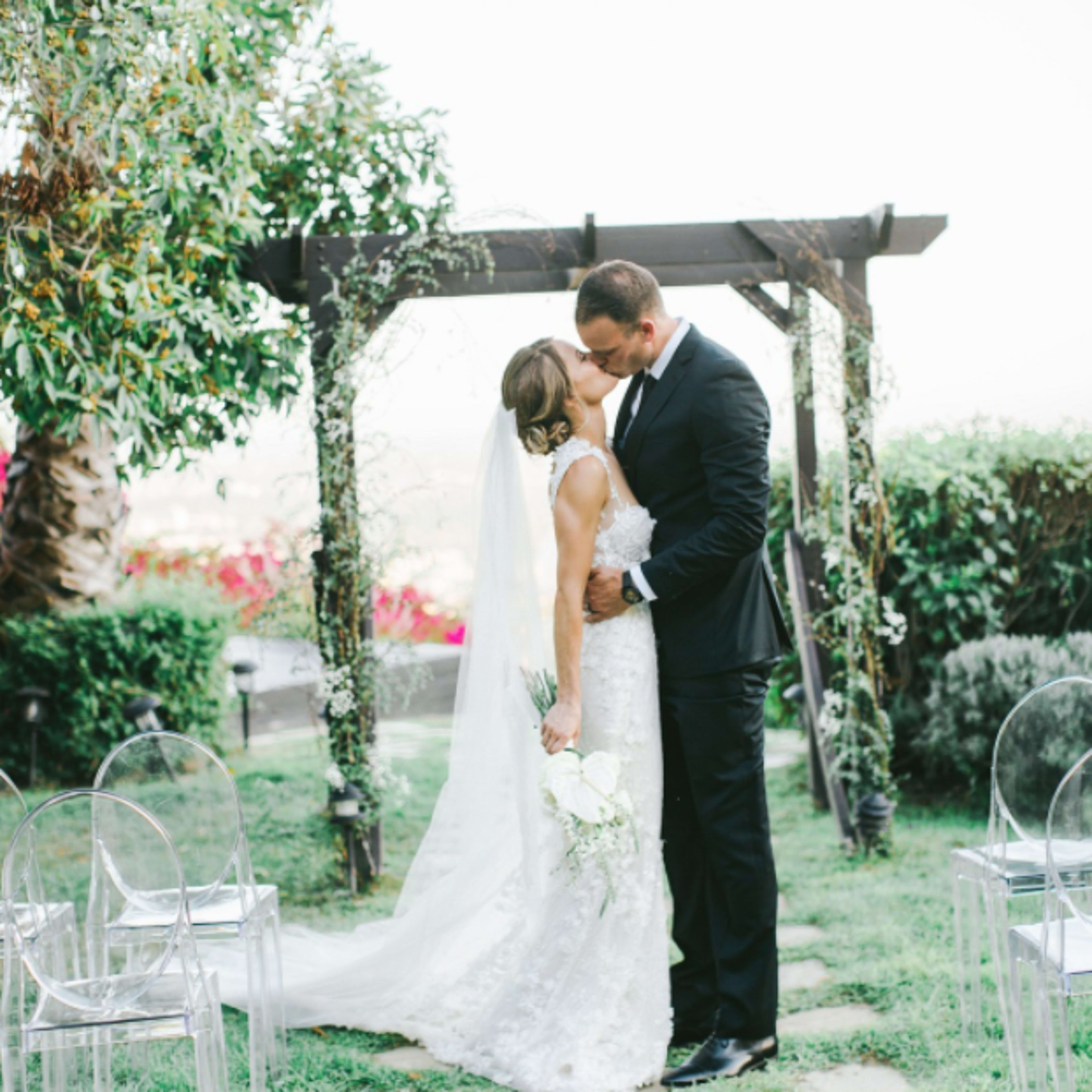 A bride and groom share a kiss under an arbor in a garden, surrounded by greenery and clear chairs arranged for guests.