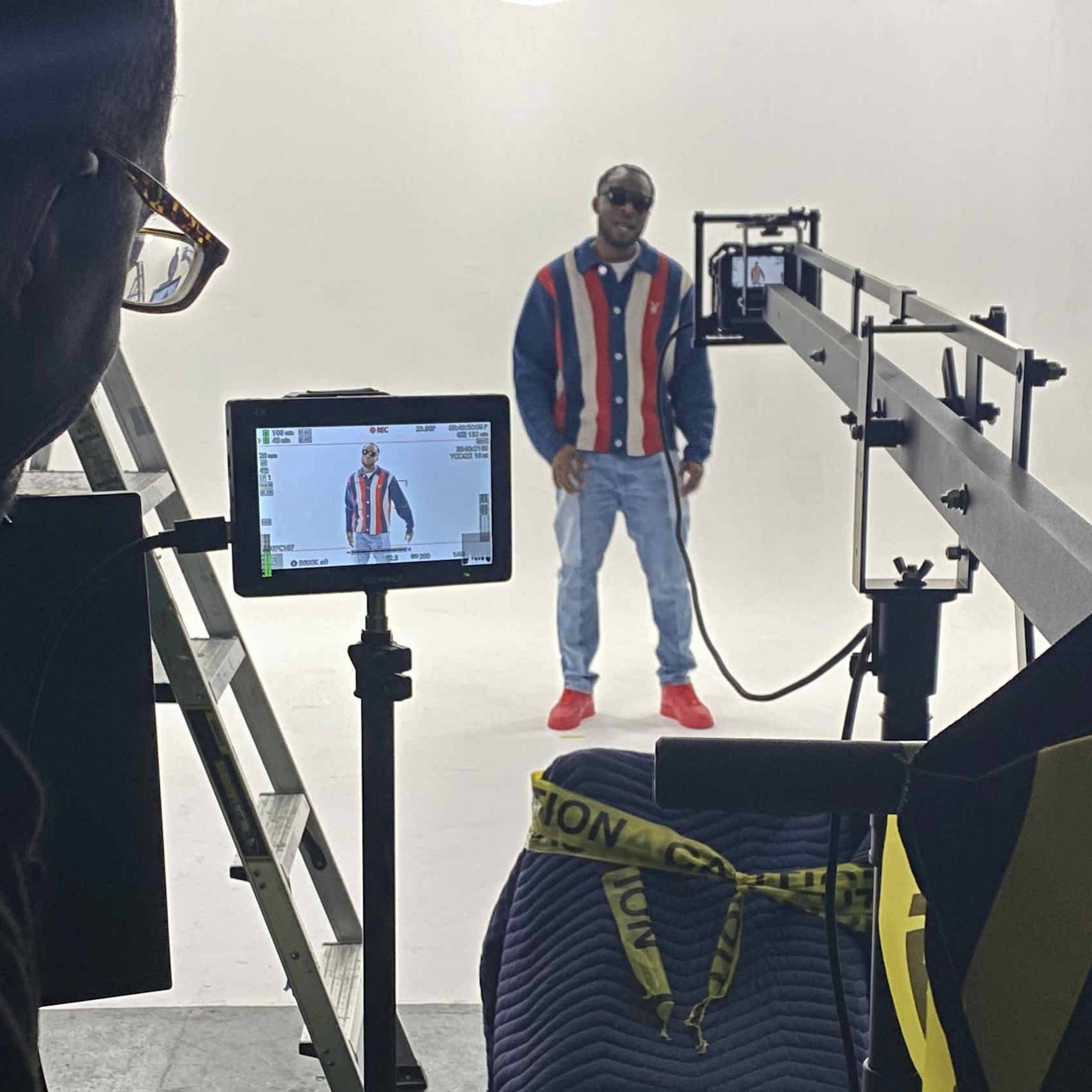 A man dressed in a striped jacket and sunglasses stands in a well-lit studio with filming equipment set up nearby.
