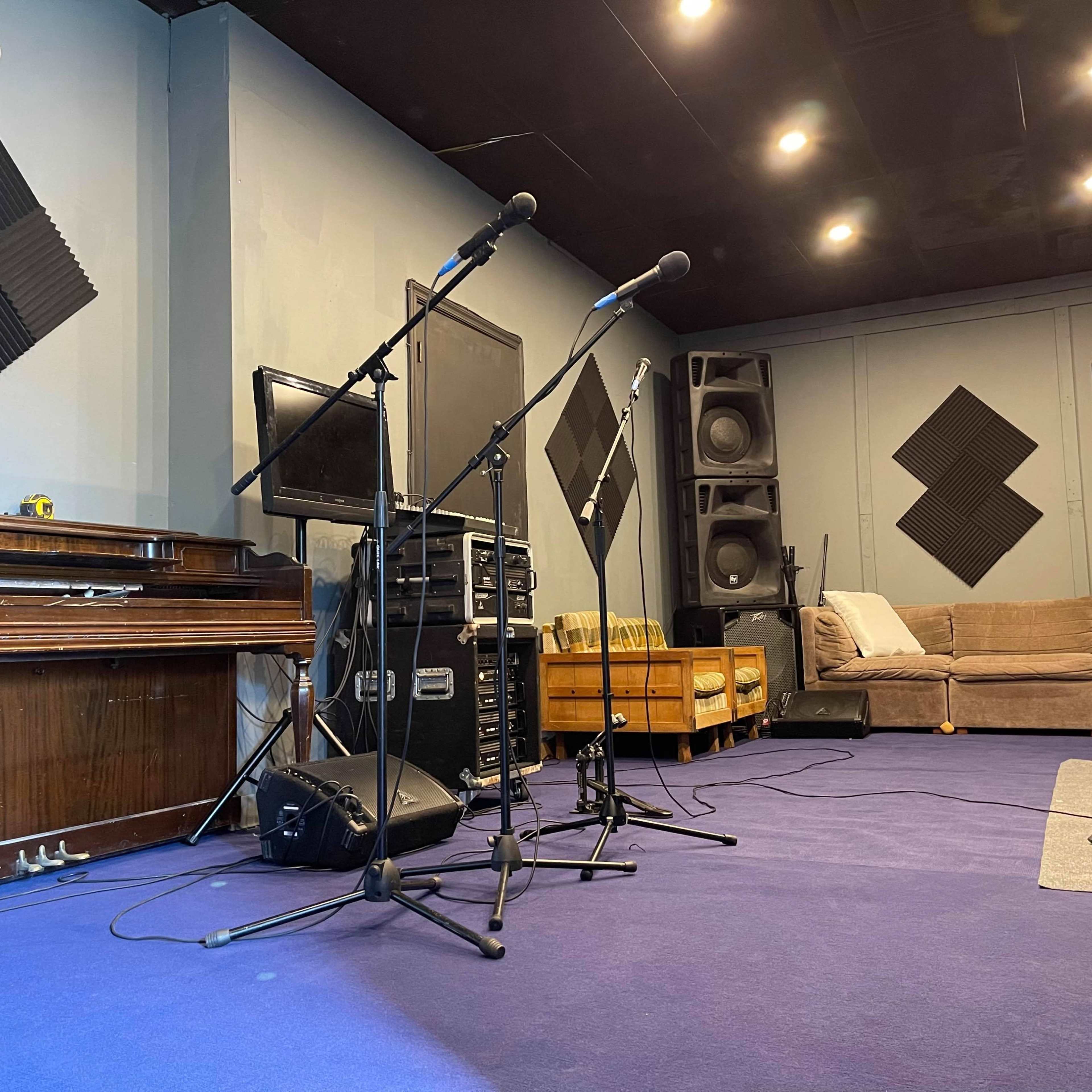 A music studio with microphones set up in front of a piano, a stack of sound equipment, and a couch in the background.