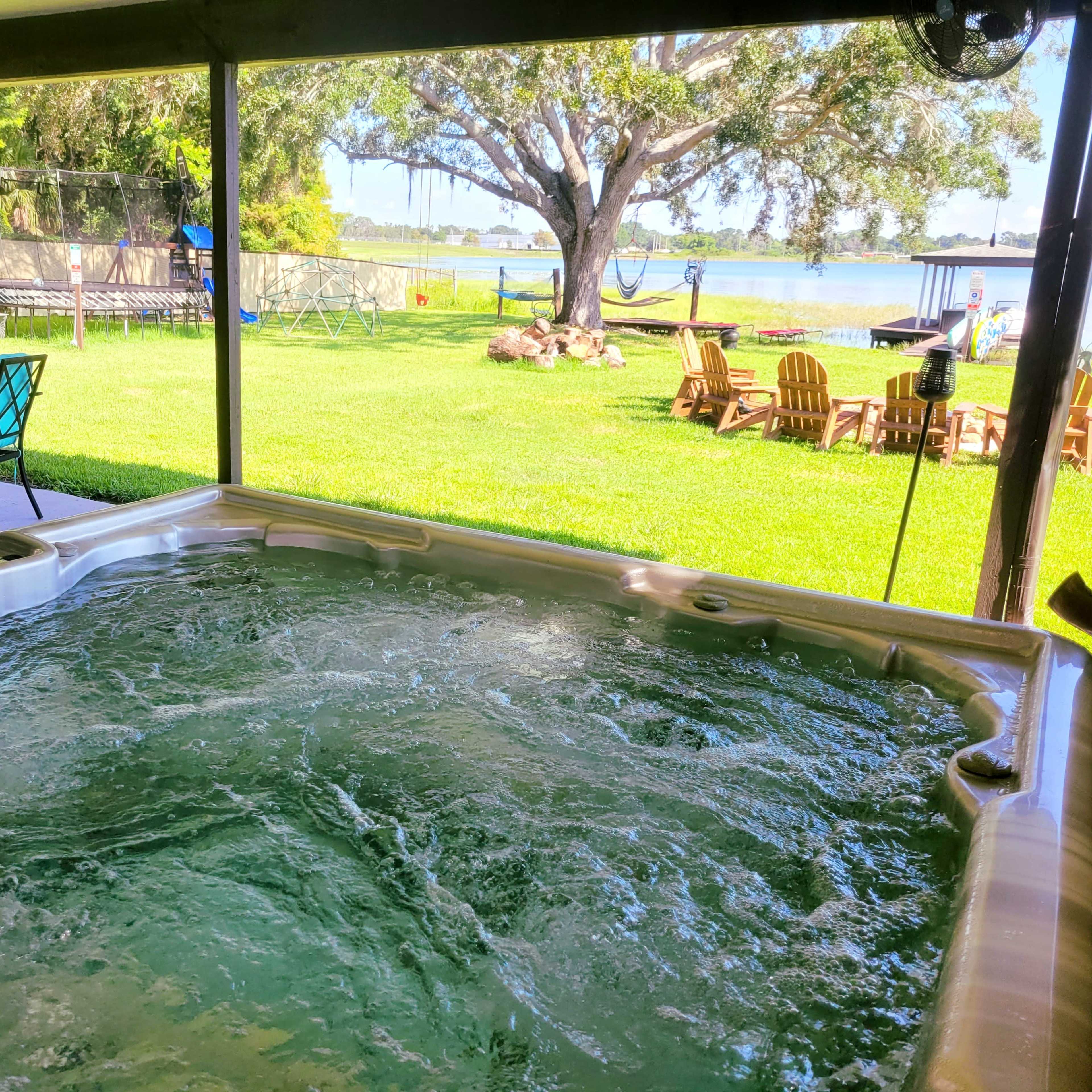 A hot tub filled with bubbling water is situated on a patio overlooking a grassy area and a lake.