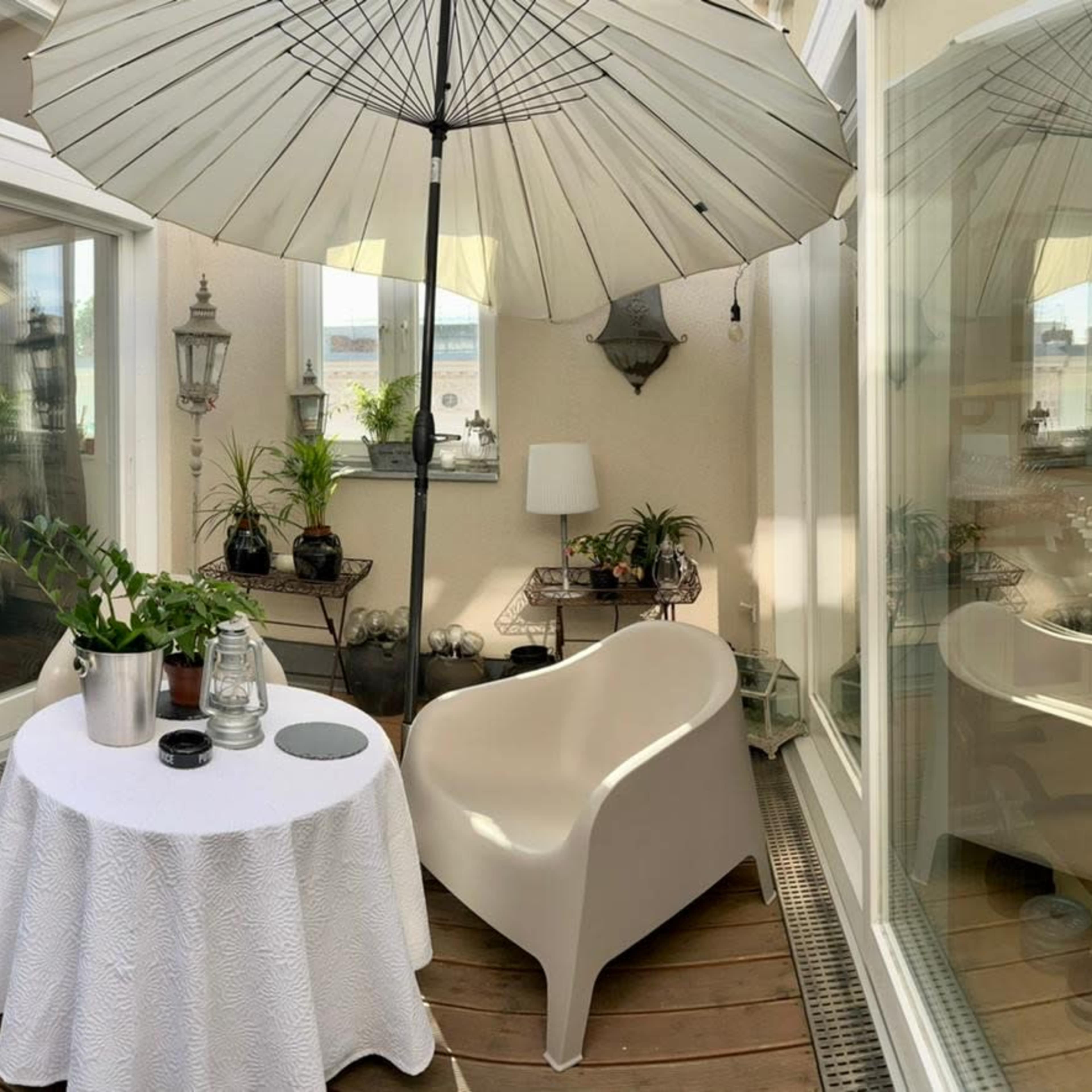 A small, enclosed balcony features a white table with a tablecloth, a chair, an umbrella, potted plants, and large windows.
