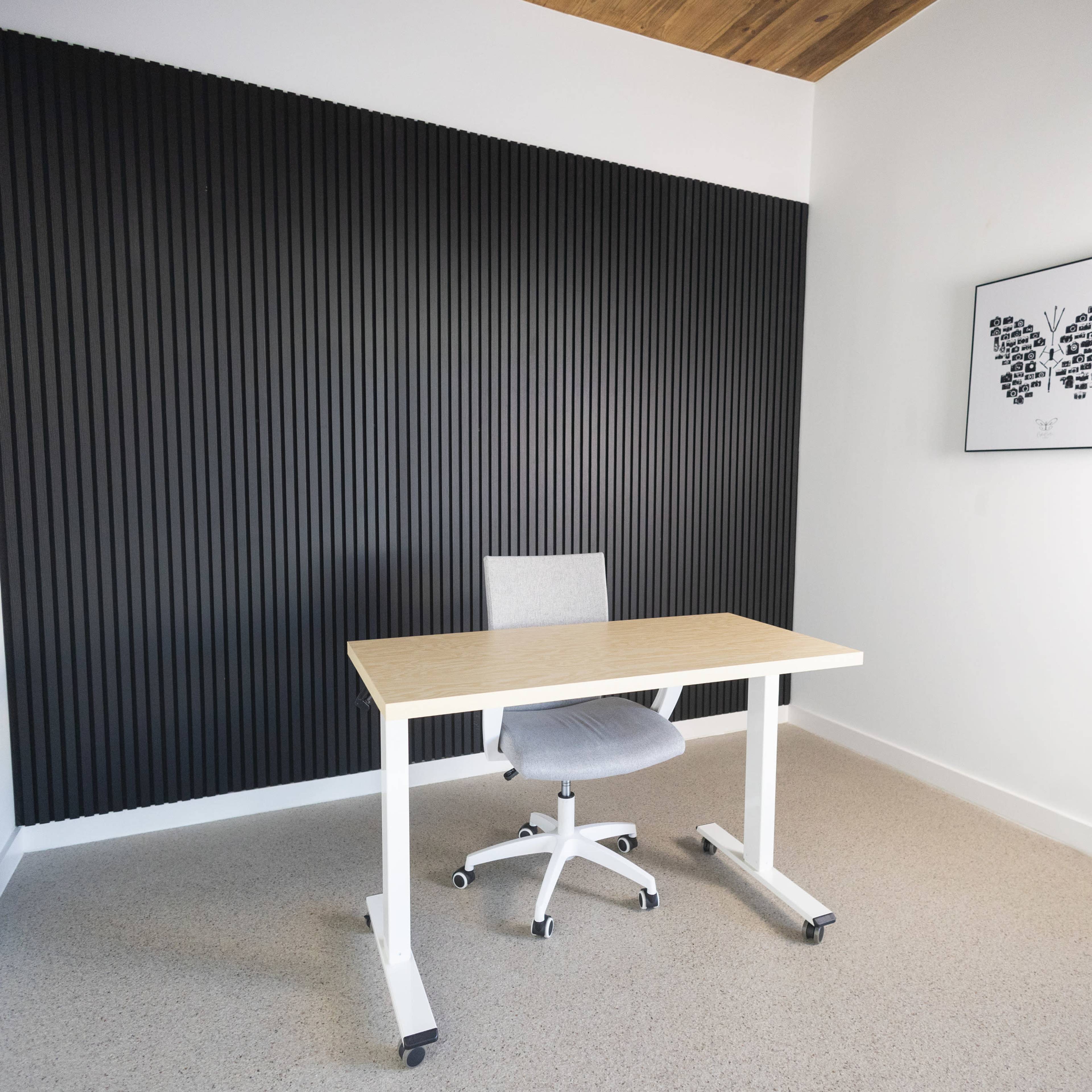 A minimalistic office space with a light wooden desk and a gray rolling chair against a black ribbed wall.
