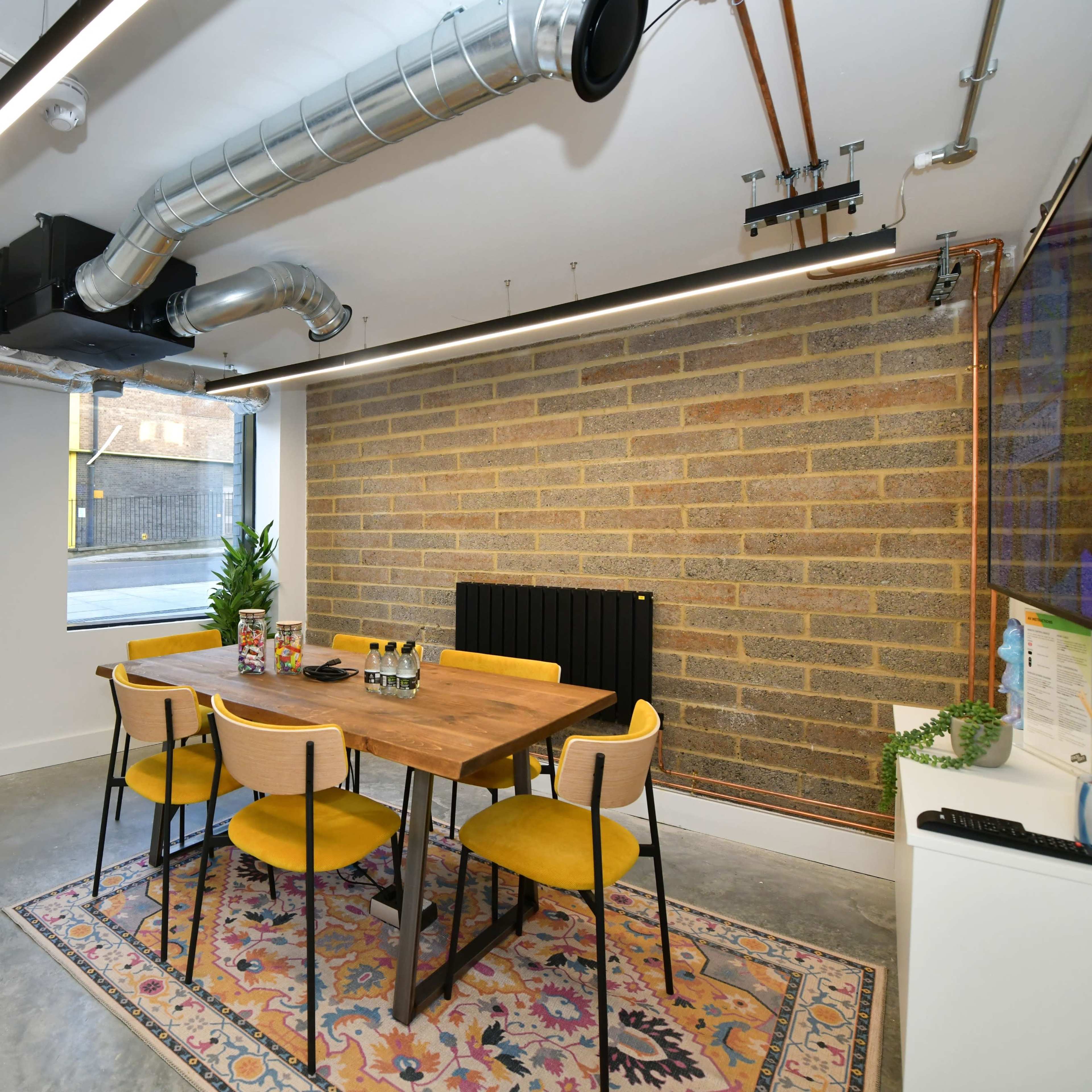 A modern dining area with a wooden table surrounded by yellow chairs, a patterned rug, and a brick wall in the background.
