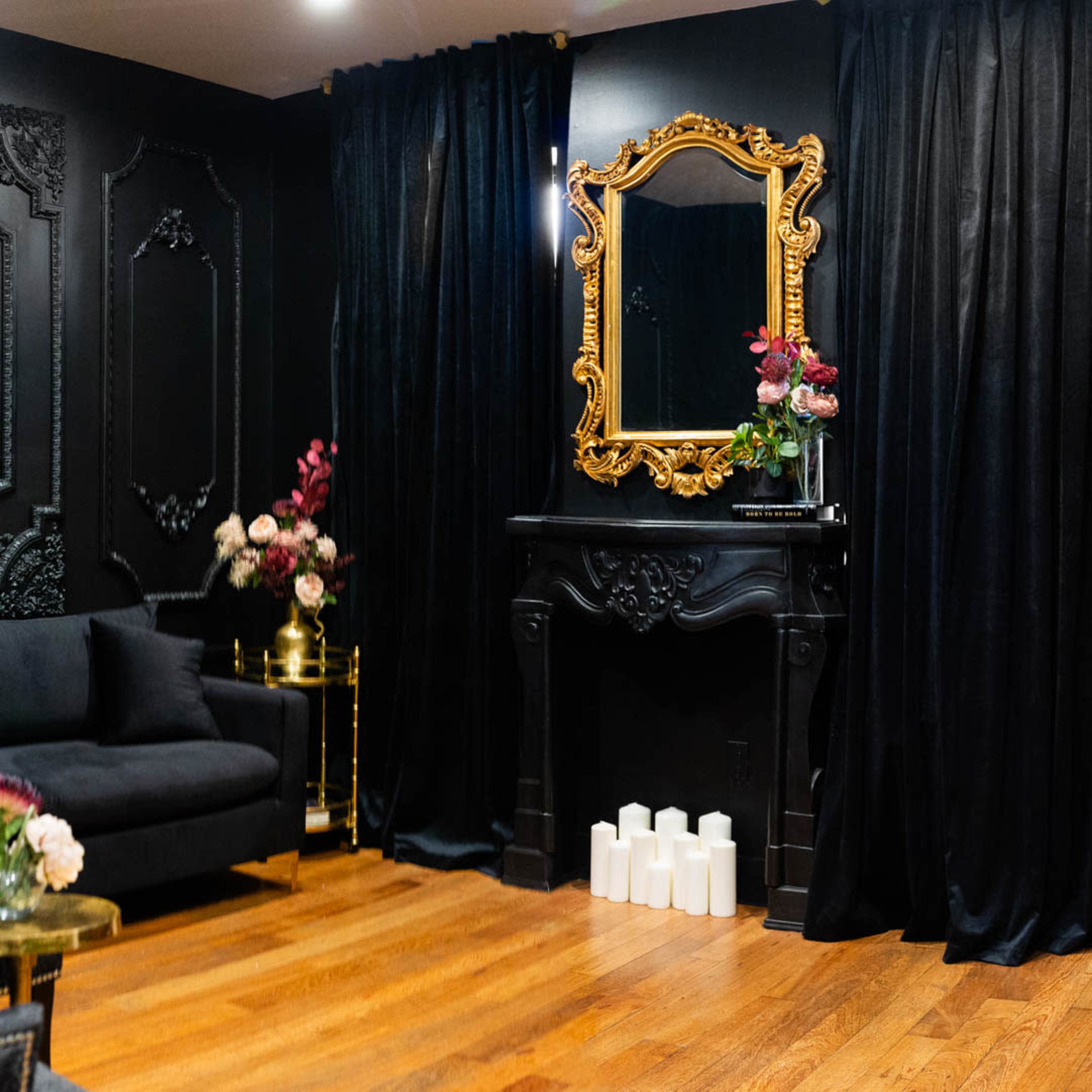 The room features a dark-walled setting with a decorative black fireplace, a large ornate gold mirror, a velvet couch, and white candles on the floor.