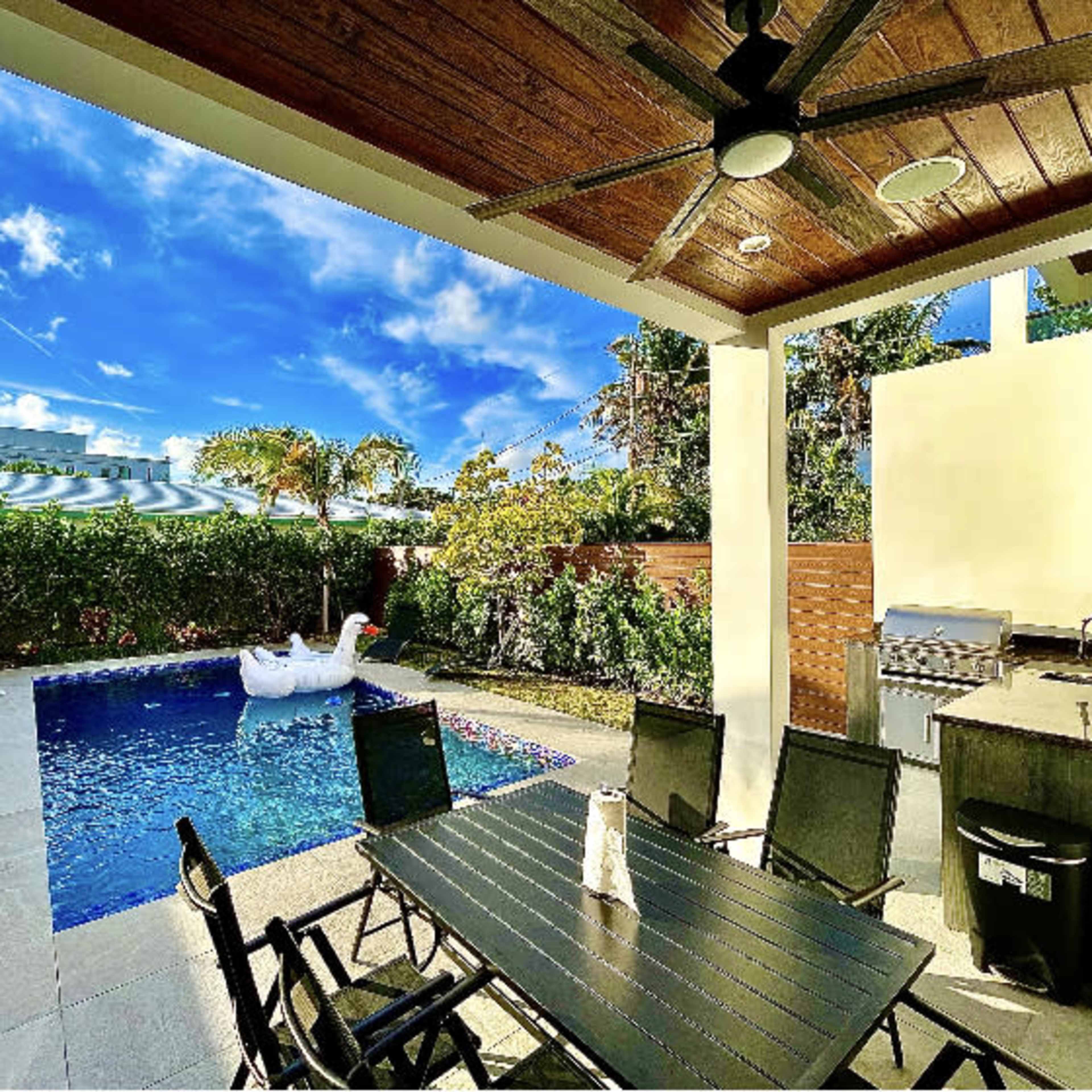A backyard patio with a dining table, a pool featuring a large inflatable swan, and lush greenery under a clear blue sky.