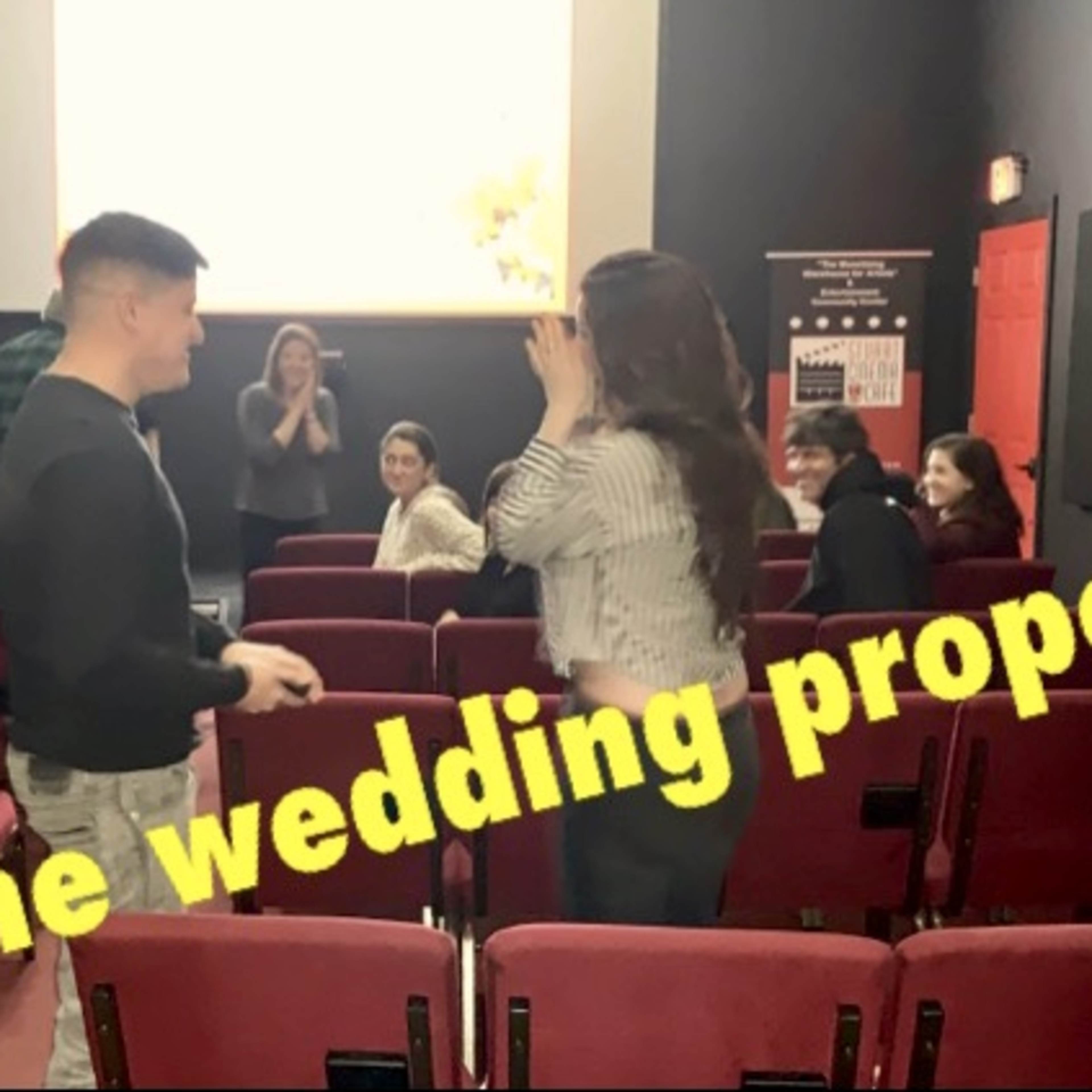 A man proposes to a woman in a theater setting while an audience watches.