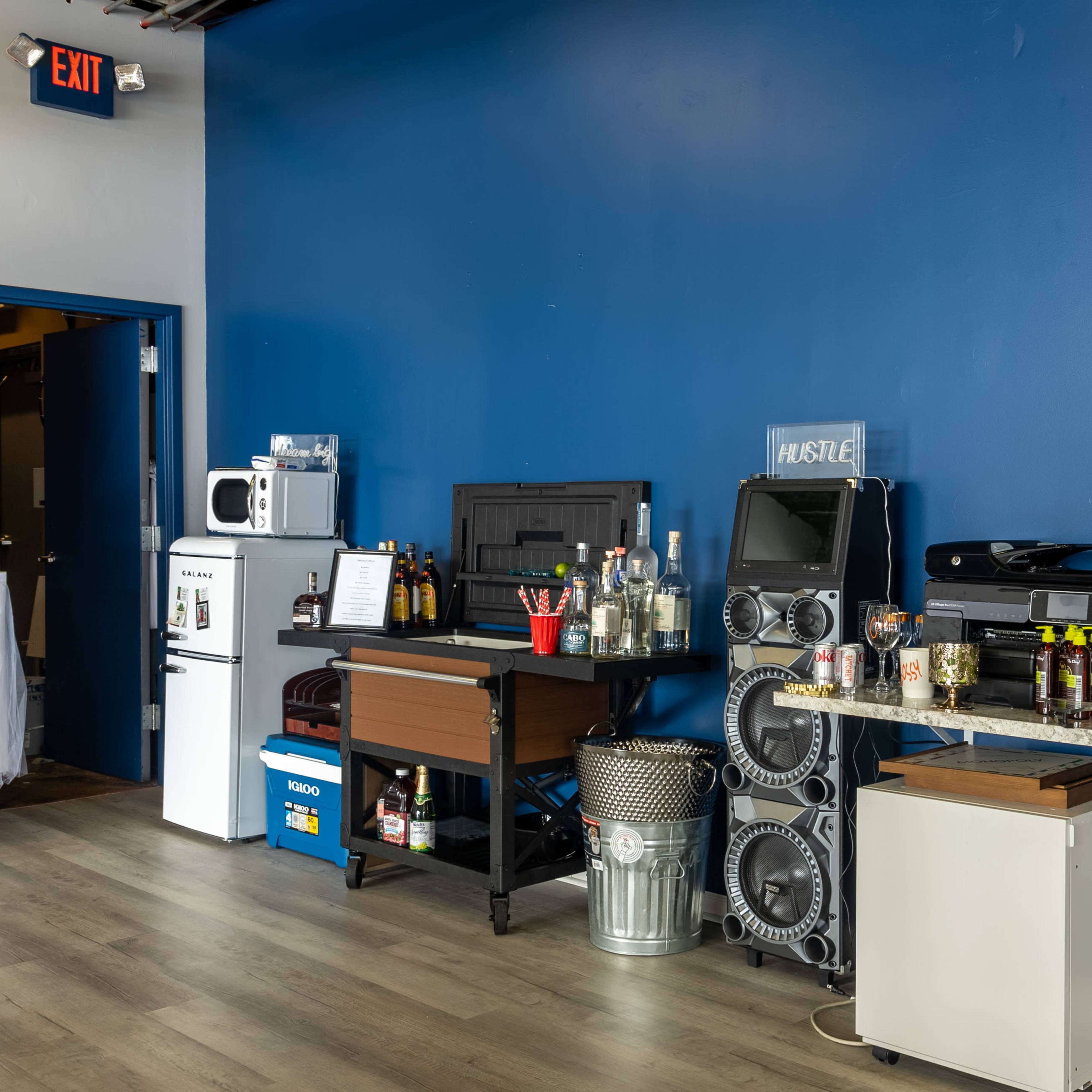 A blue-walled room with a variety of appliances and items on counters, including a microwave, a speaker system, and bins filled with various containers.