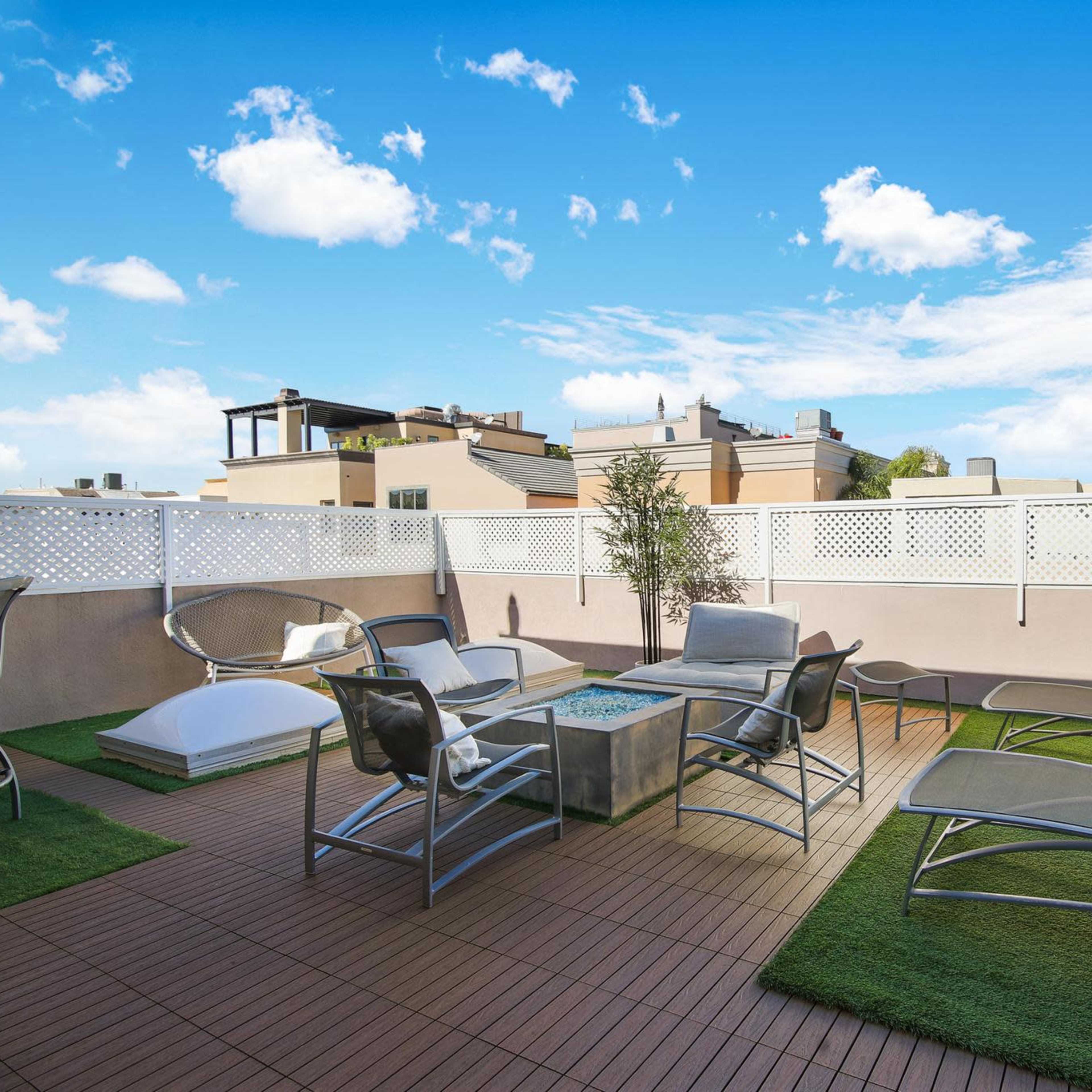 A modern rooftop terrace featuring lounge chairs, a small table, and artificial grass under a bright blue sky.