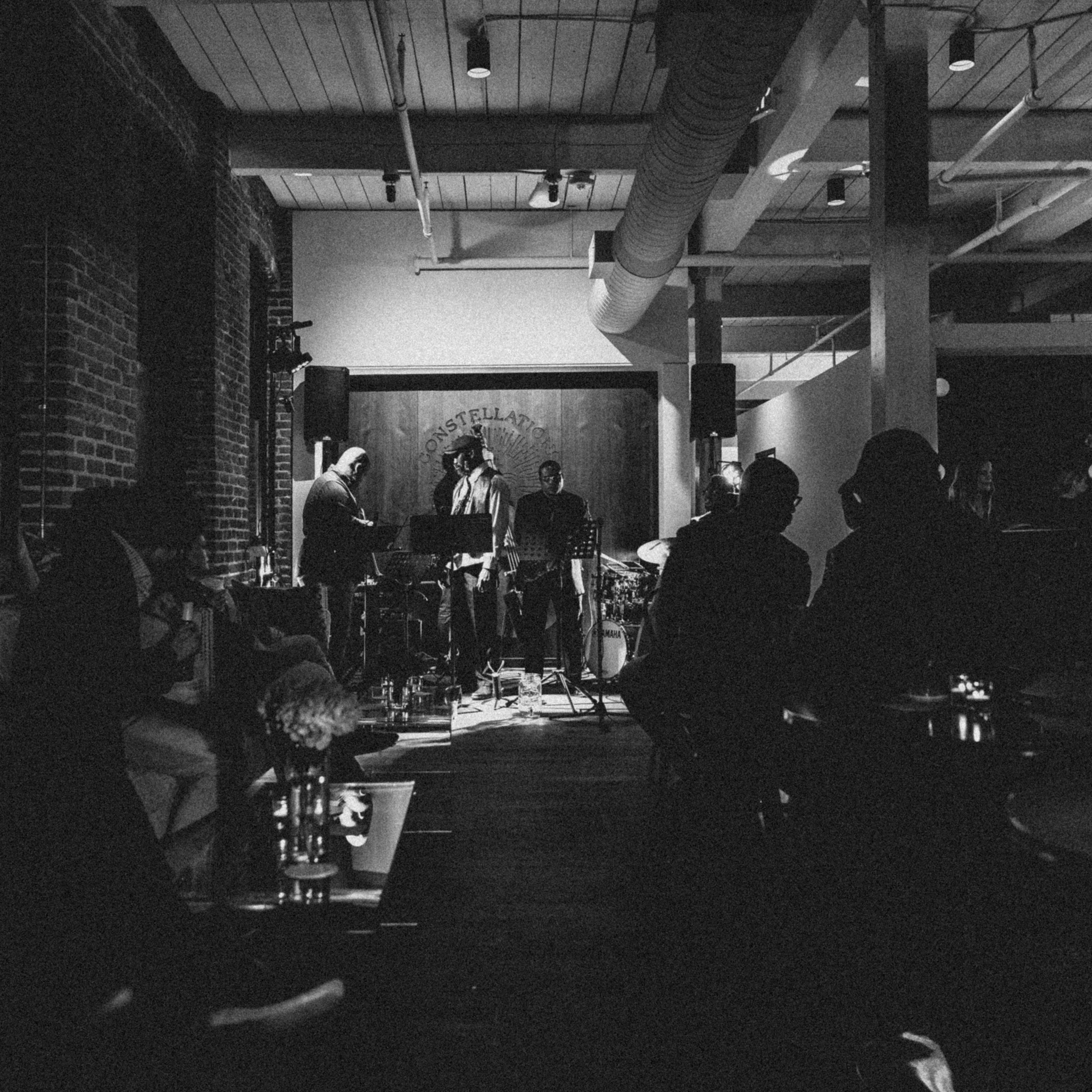 A jazz band performs on a dimly lit stage in a brick-walled venue while an audience listens intently.