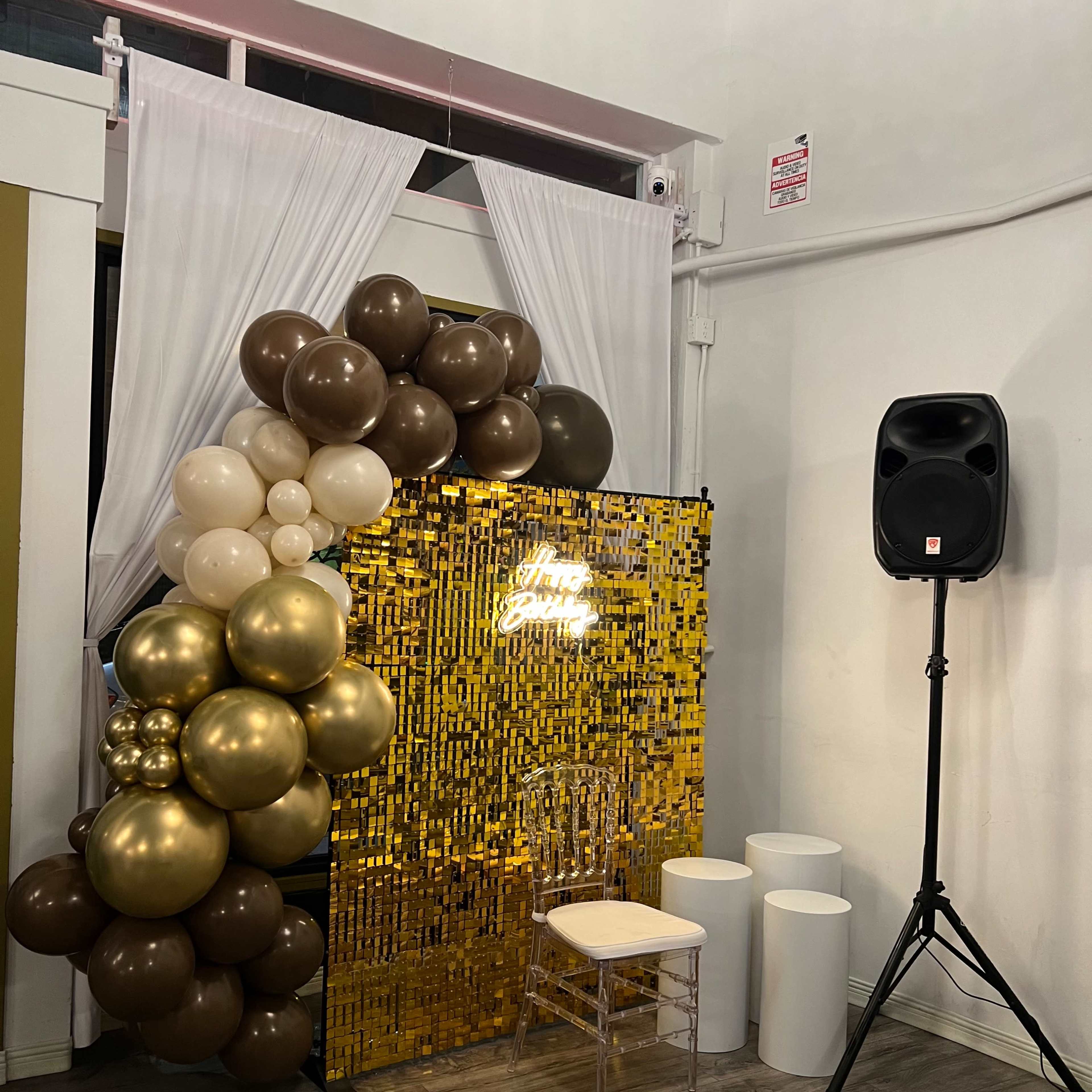 A decorated corner featuring a gold and white balloon backdrop, an illuminated sign, a clear acrylic chair, and a speaker on a stand.