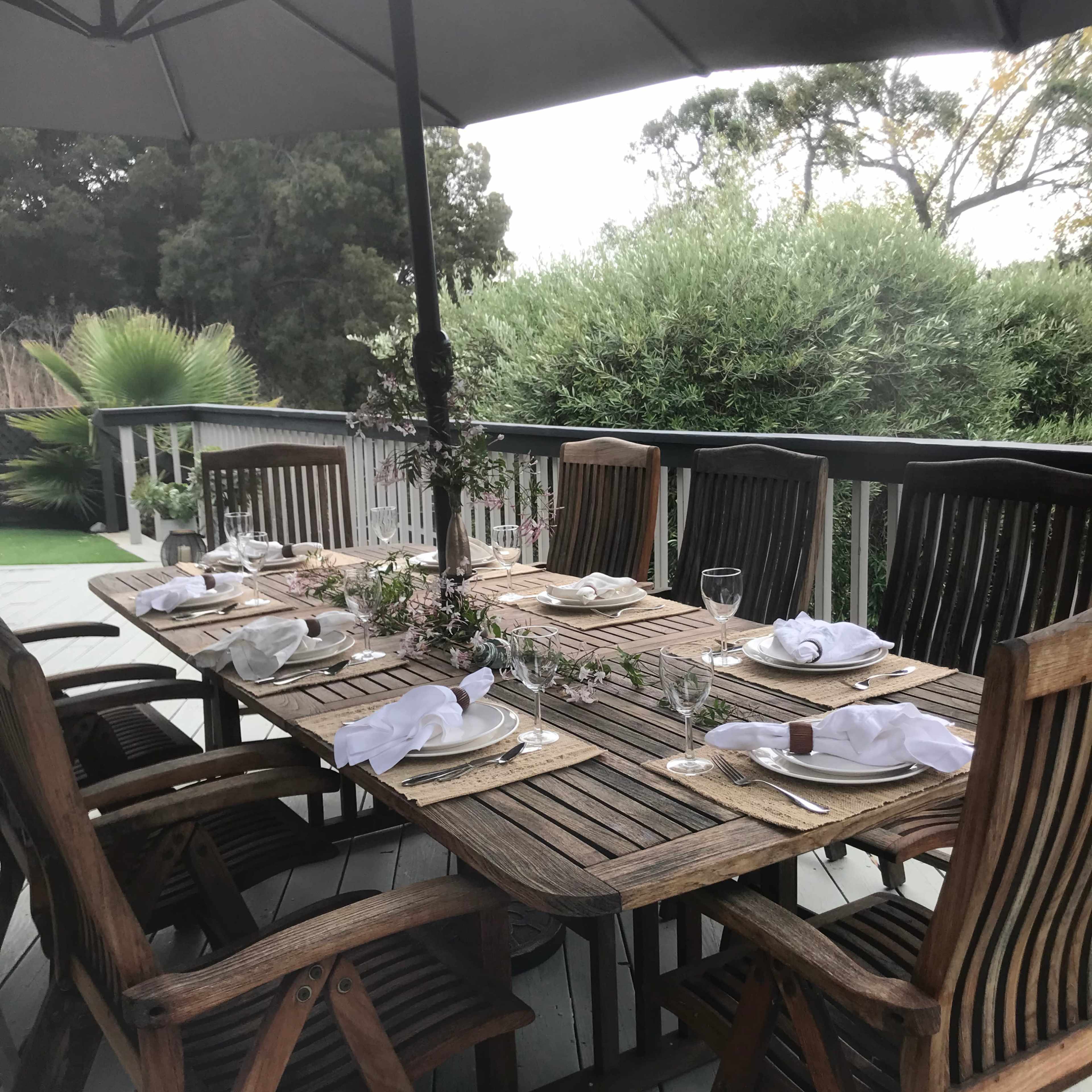 A wooden dining table is set for a meal, complete with plates, utensils, and glasses, under an umbrella on a deck surrounded by greenery.