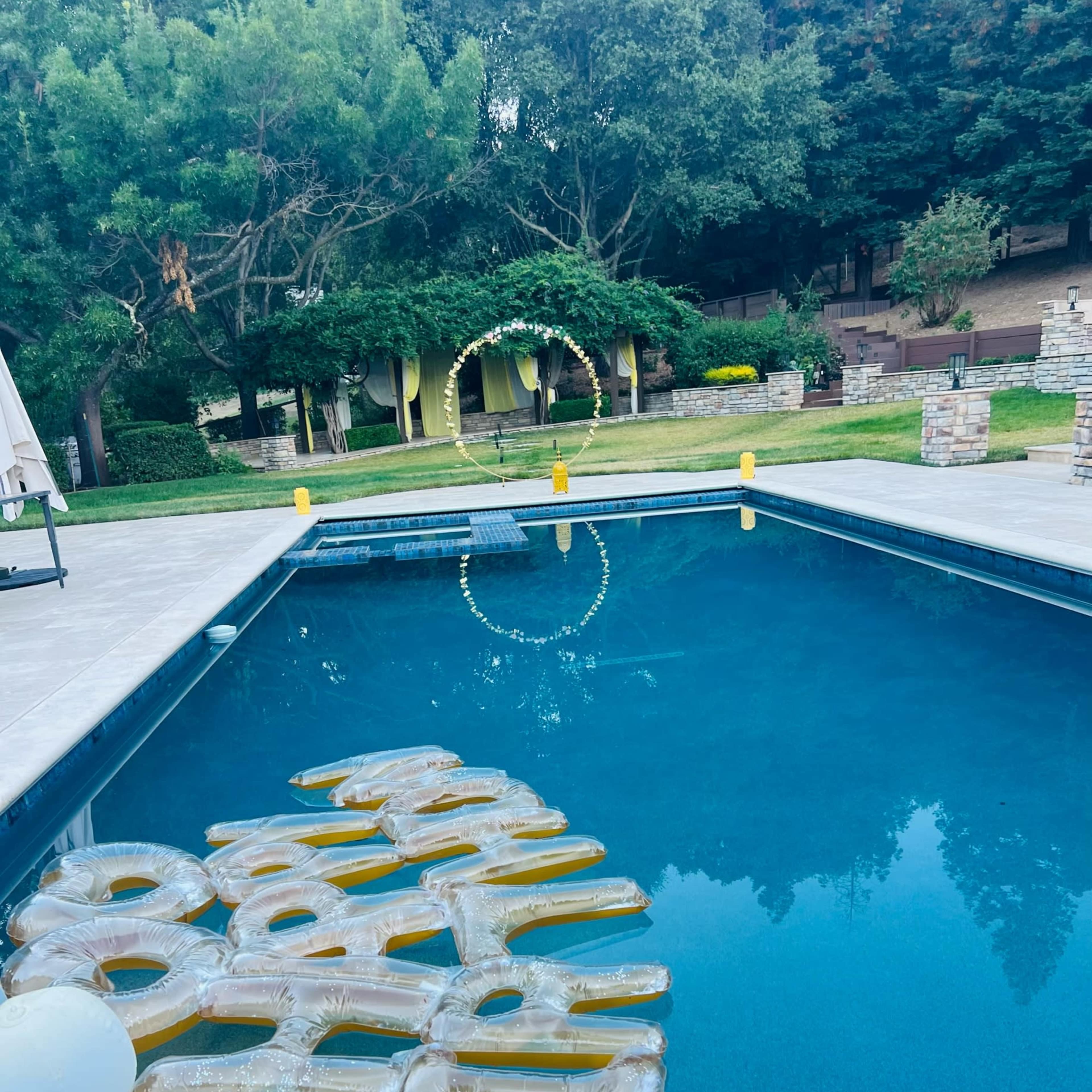 A swimming pool with oversized, transparent letters floating on the water, surrounded by greenery and a decorative arch in the background.