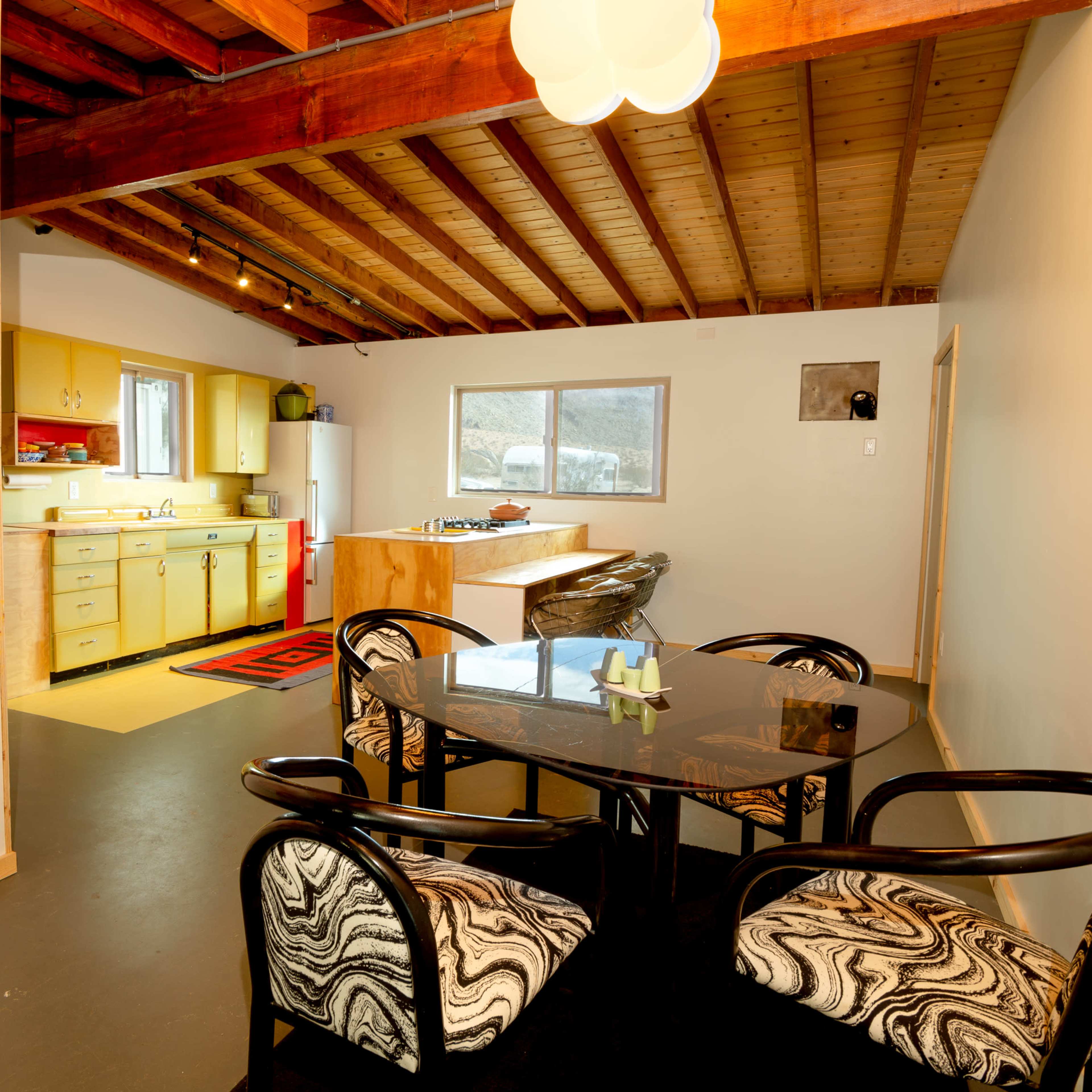 A dining area features a black table with patterned chairs and adjoins a kitchen with yellow cabinets and a window.