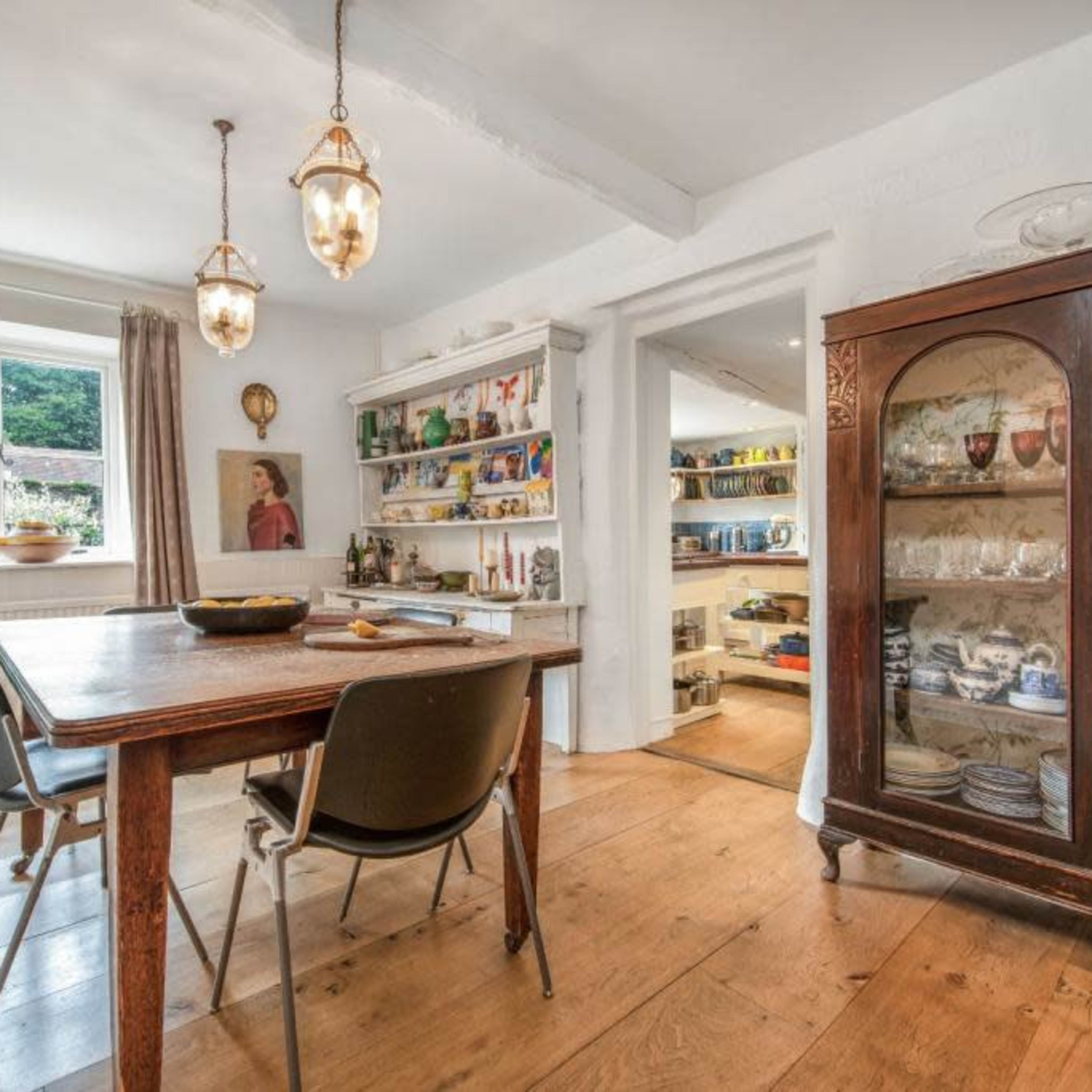 A wooden dining table with three chairs is situated in a well-lit room featuring a glass display cabinet and shelves filled with colorful dishes and artwork.