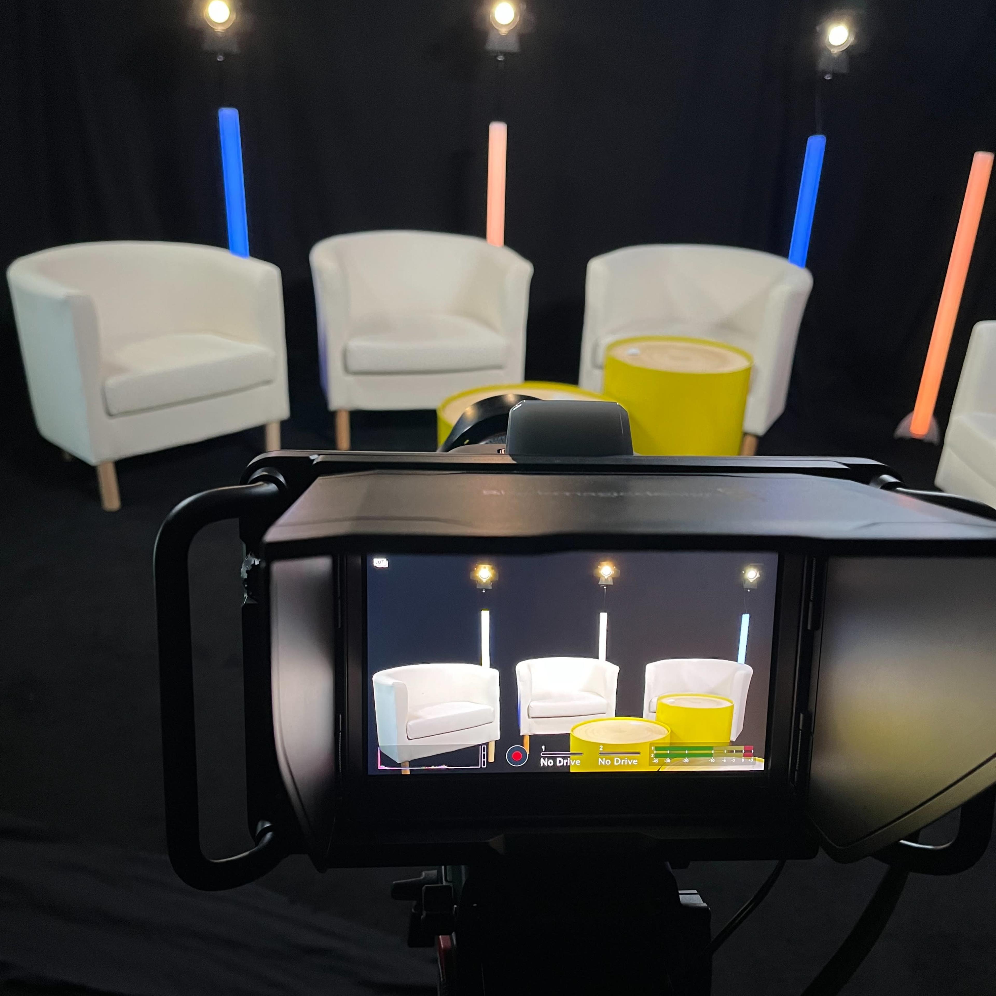 A camera is positioned to capture a stage setup featuring two white chairs and a small yellow table, with lights in the background.