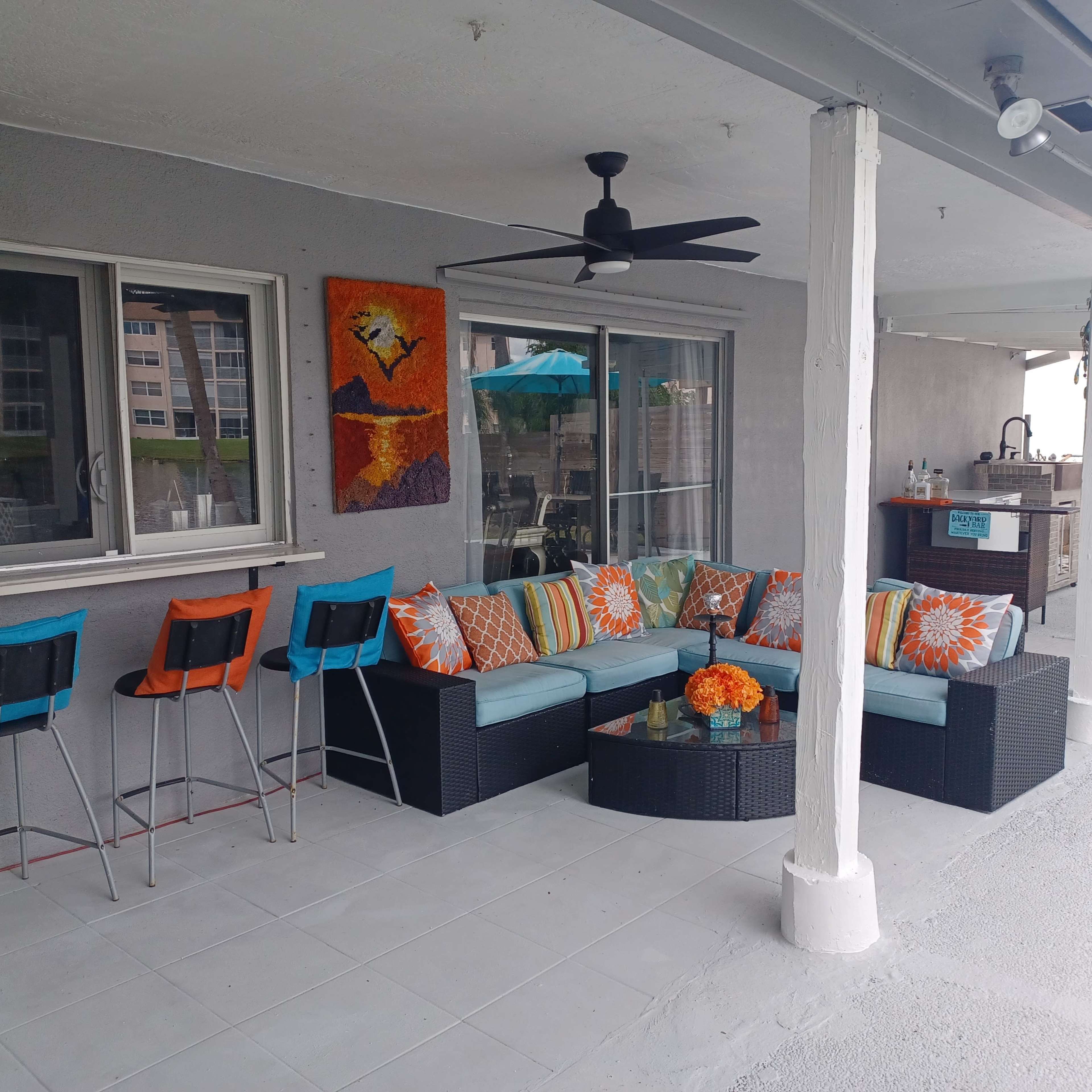 A covered patio features a wicker seating area adorned with colorful cushions, alongside a bar with stools and a backdrop of a beachside view.