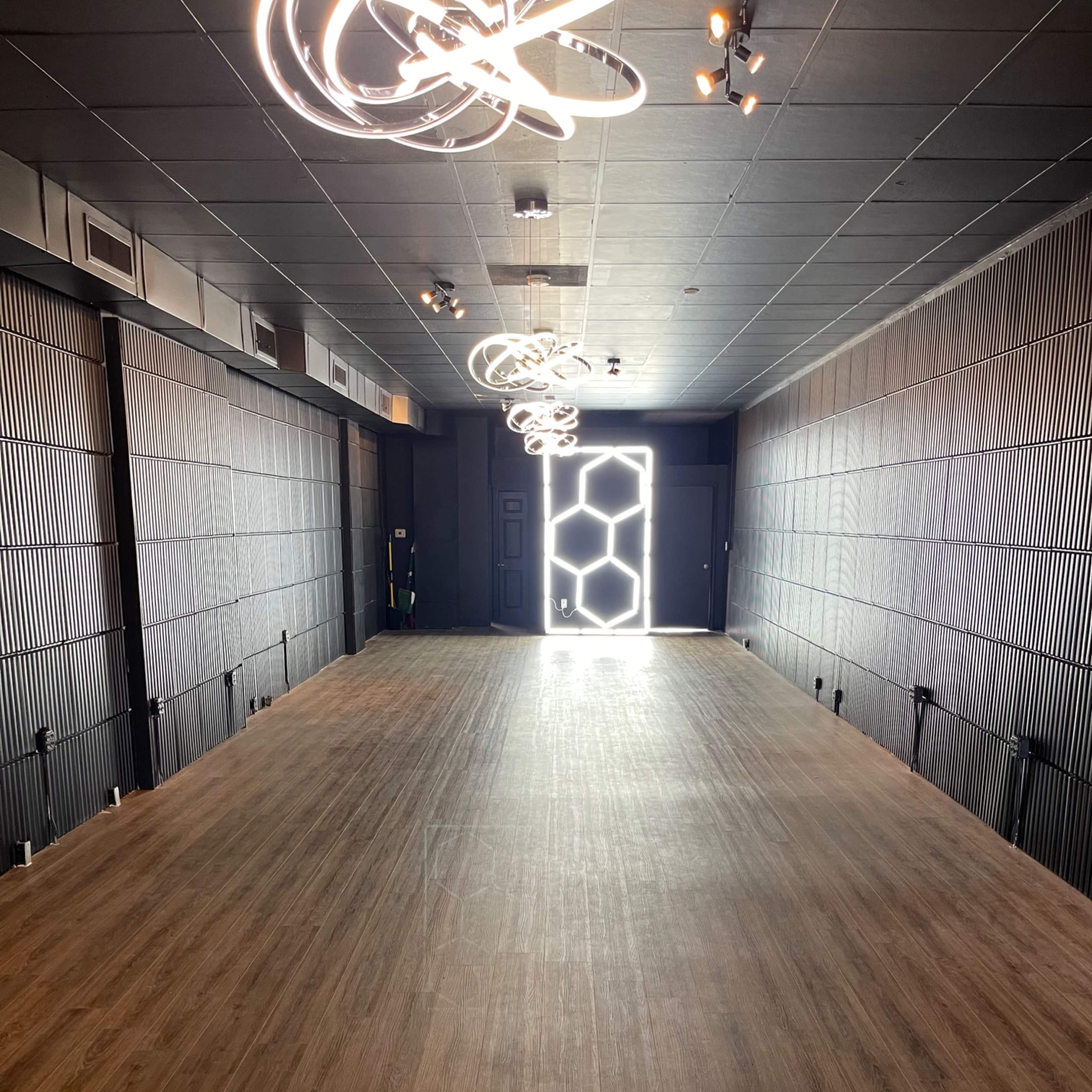 A spacious, well-lit room with wooden flooring and modern light fixtures, leading to a door at the far end.