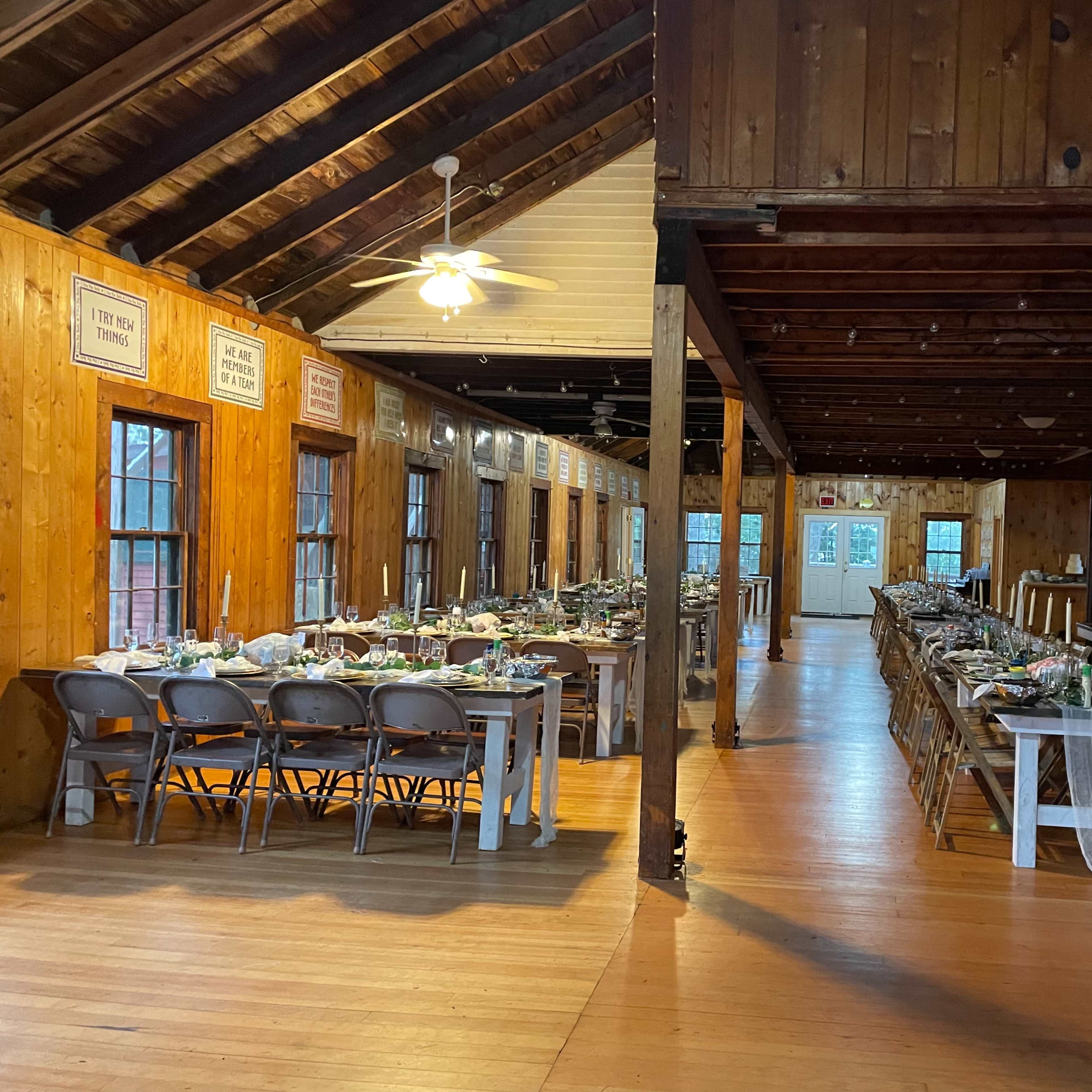 A wooden hall set up for a dining event, featuring neatly arranged tables with floral centerpieces and chairs, under a slatted wooden ceiling.