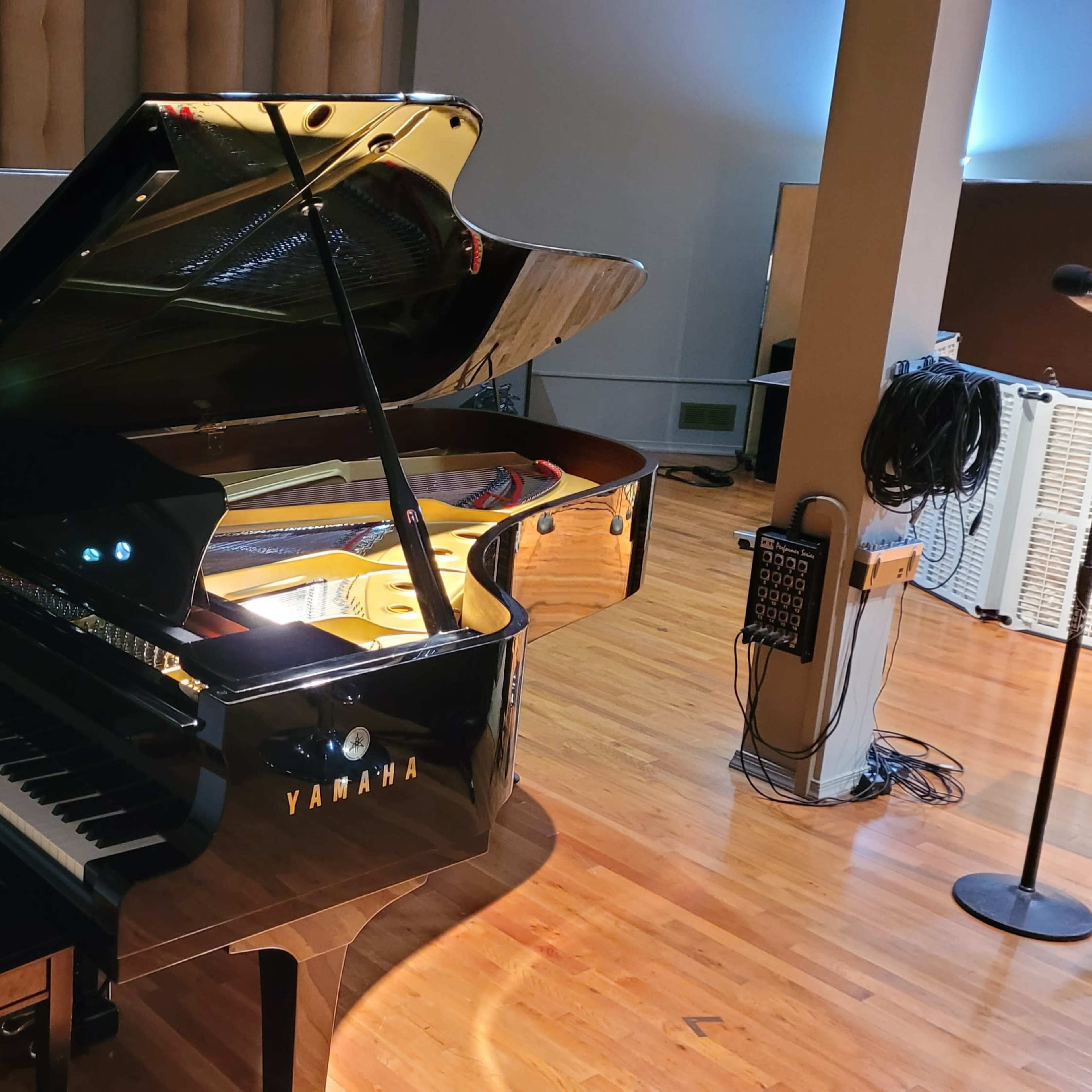 A black Yamaha grand piano is positioned in a music studio, accompanied by a keyboard and music stand in the background.