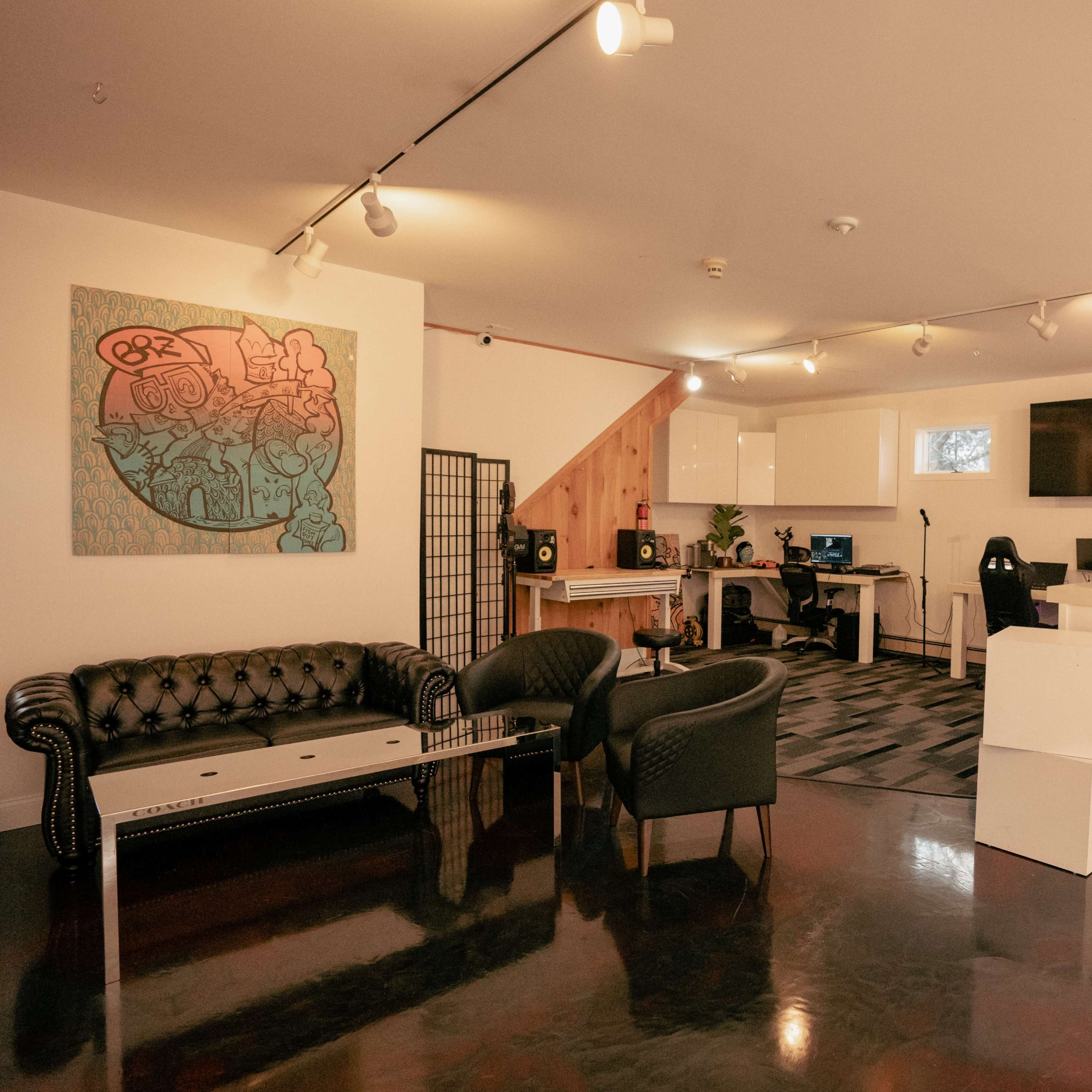 A modern, well-lit studio space with a black leather couch, two chairs, a white reception desk, and a large screen on the wall, while colorful artwork adorns one side.