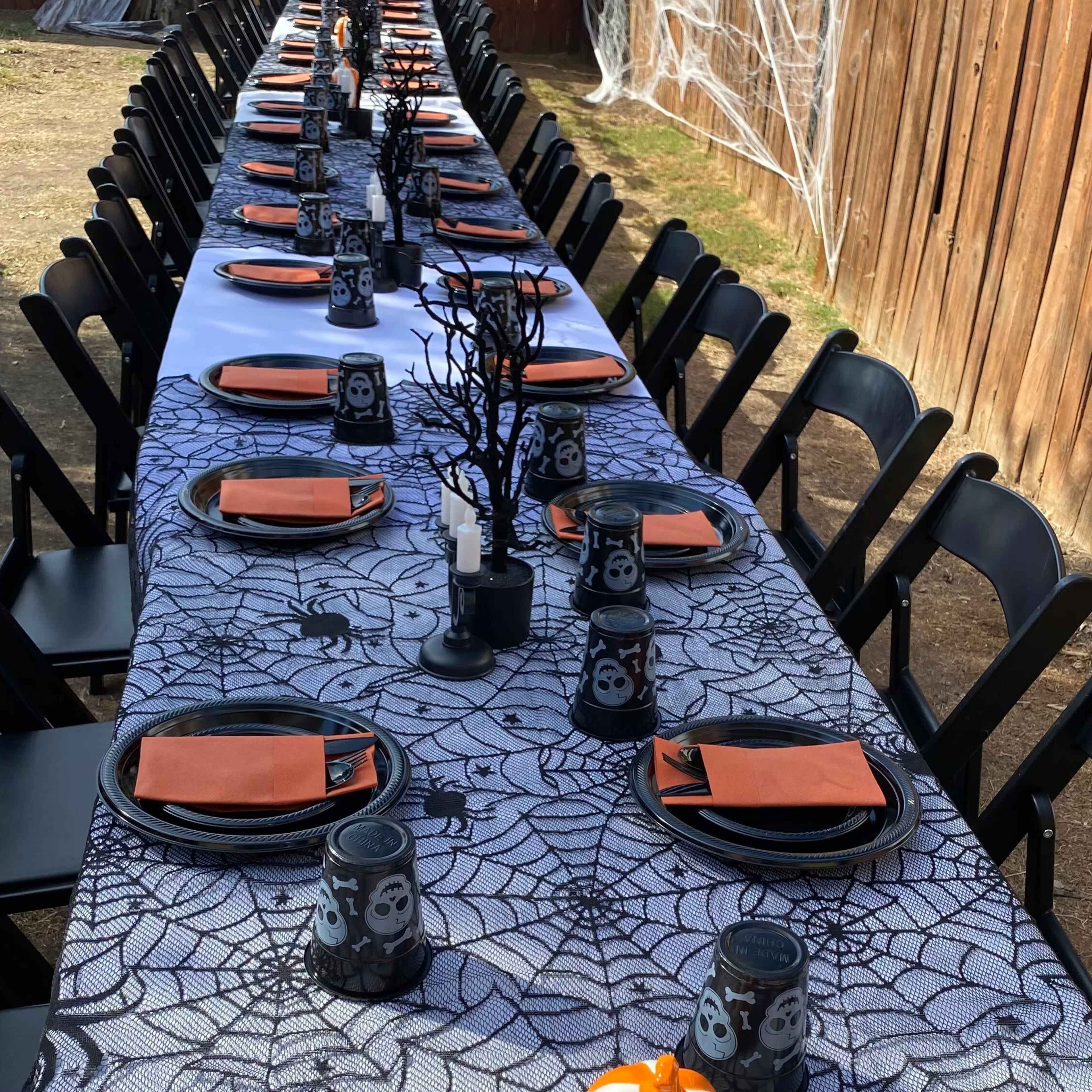 A long table is set outdoors with a spiderweb tablecloth, black chairs, and candles, featuring orange plates and spooky-themed decorations.