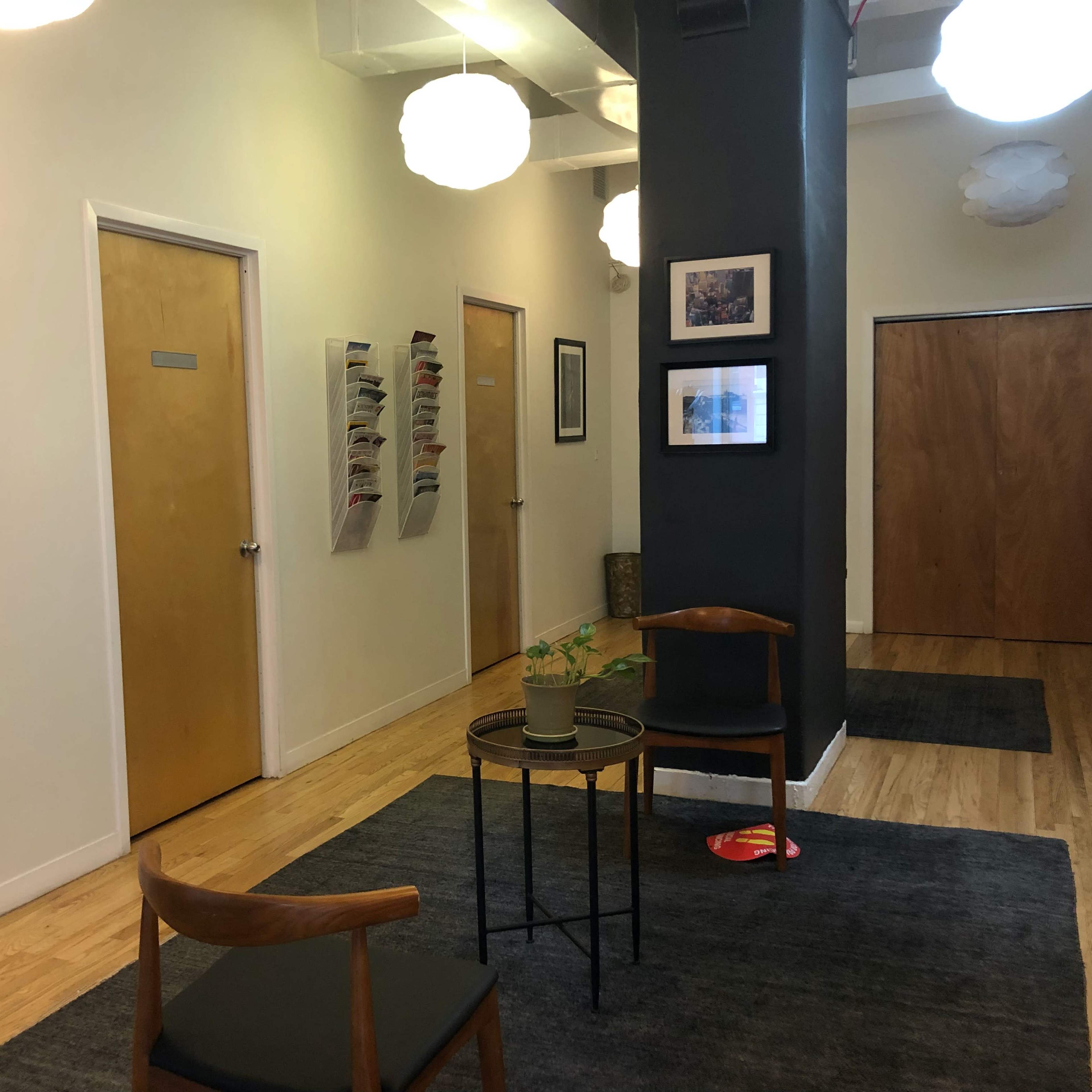 A modern hallway with wooden floors, two closed doors, a framed floral photo on the wall, a circular table with a small plant, and two chairs.