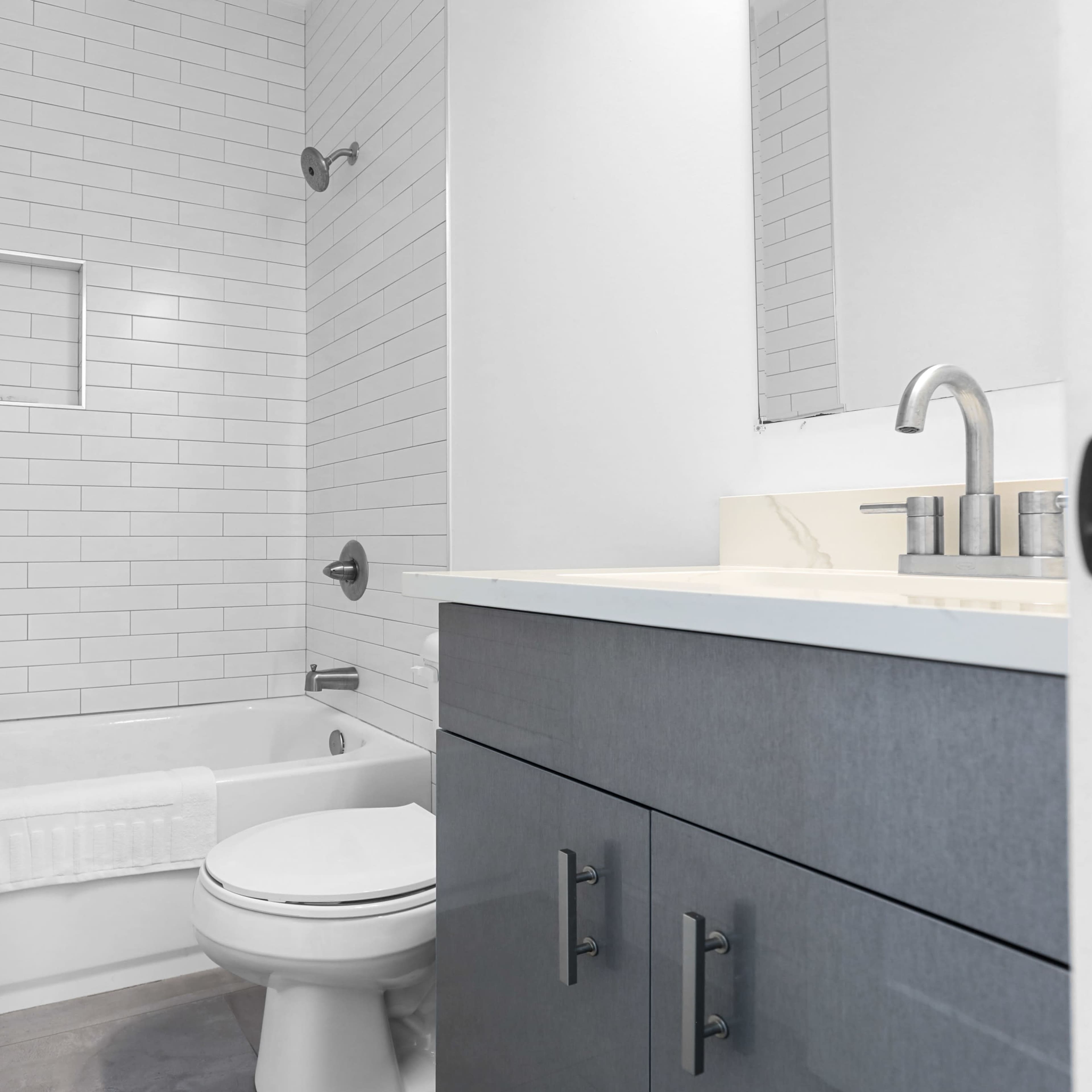 A clean bathroom featuring white subway tile walls, a bathtub with a shower, a toilet, and a dark gray vanity with a countertop.